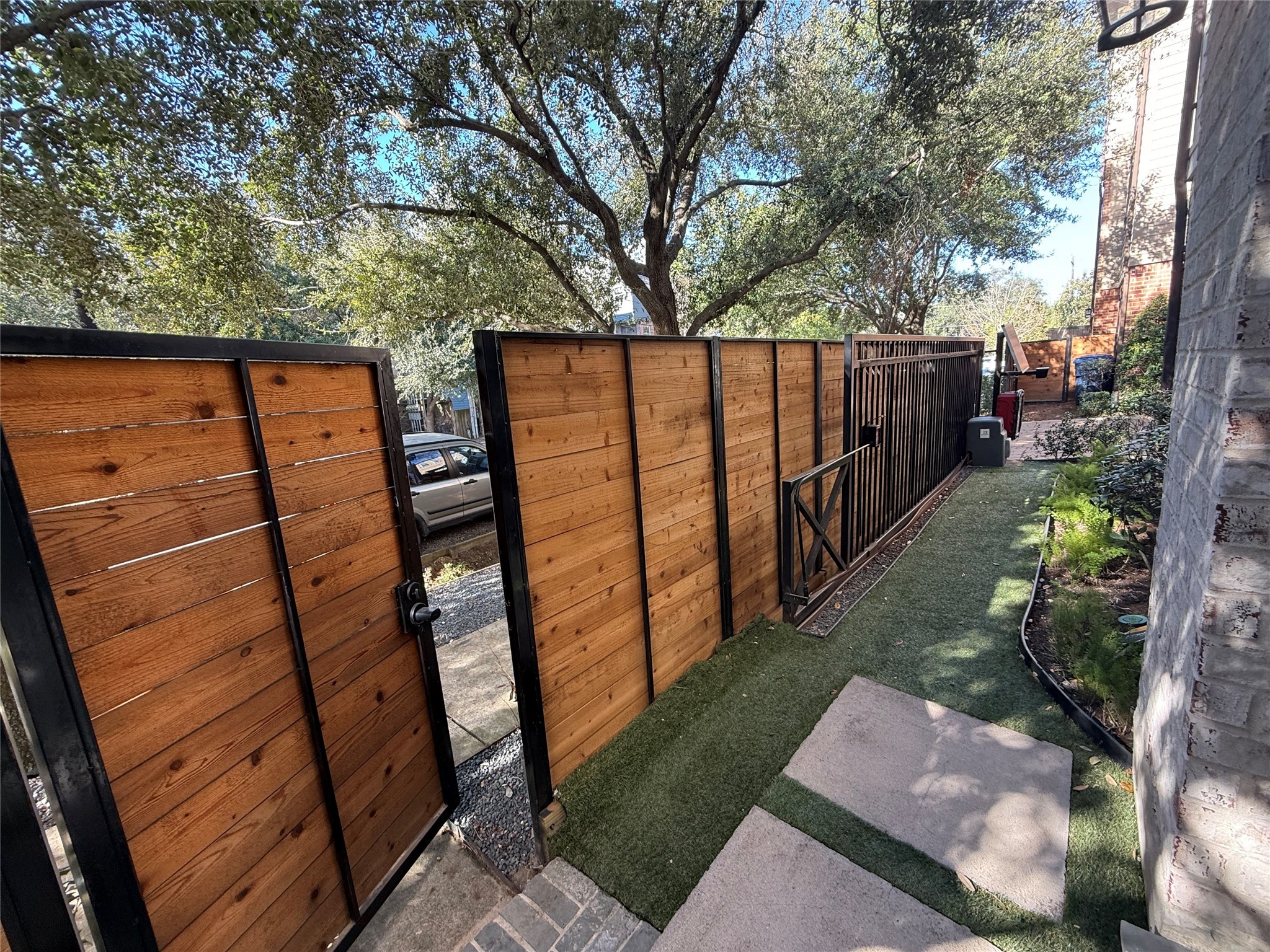 620 Rutland Street, Unit A Houston, TX 77007 - Photo 6 of 46 This photo shows a side yard with a wooden fence and gate, offering privacy and security. The area features a narrow path with artificial turf and is lined with small plants. Overhead, mature trees provide shade and a tranquil atmosphere.