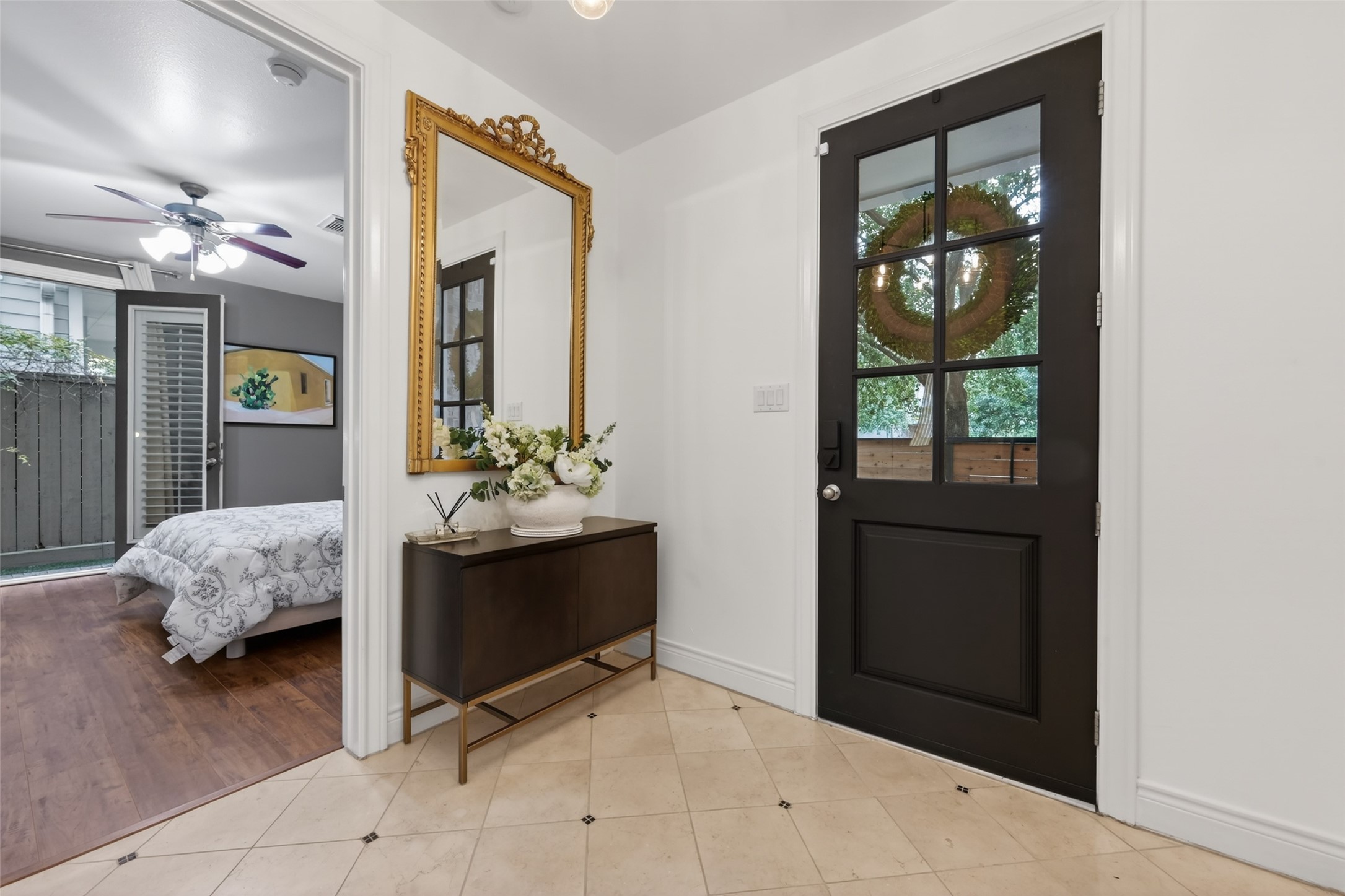 620 Rutland Street, Unit A Houston, TX 77007 - Photo 7 of 46 This photo showcases a stylish entryway with a black door featuring a large window, and a gold-framed mirror above a modern console table. Adjacent is a secondary bedroom with new wood flooring, a ceiling fan, and access to the wrap around side yard.
