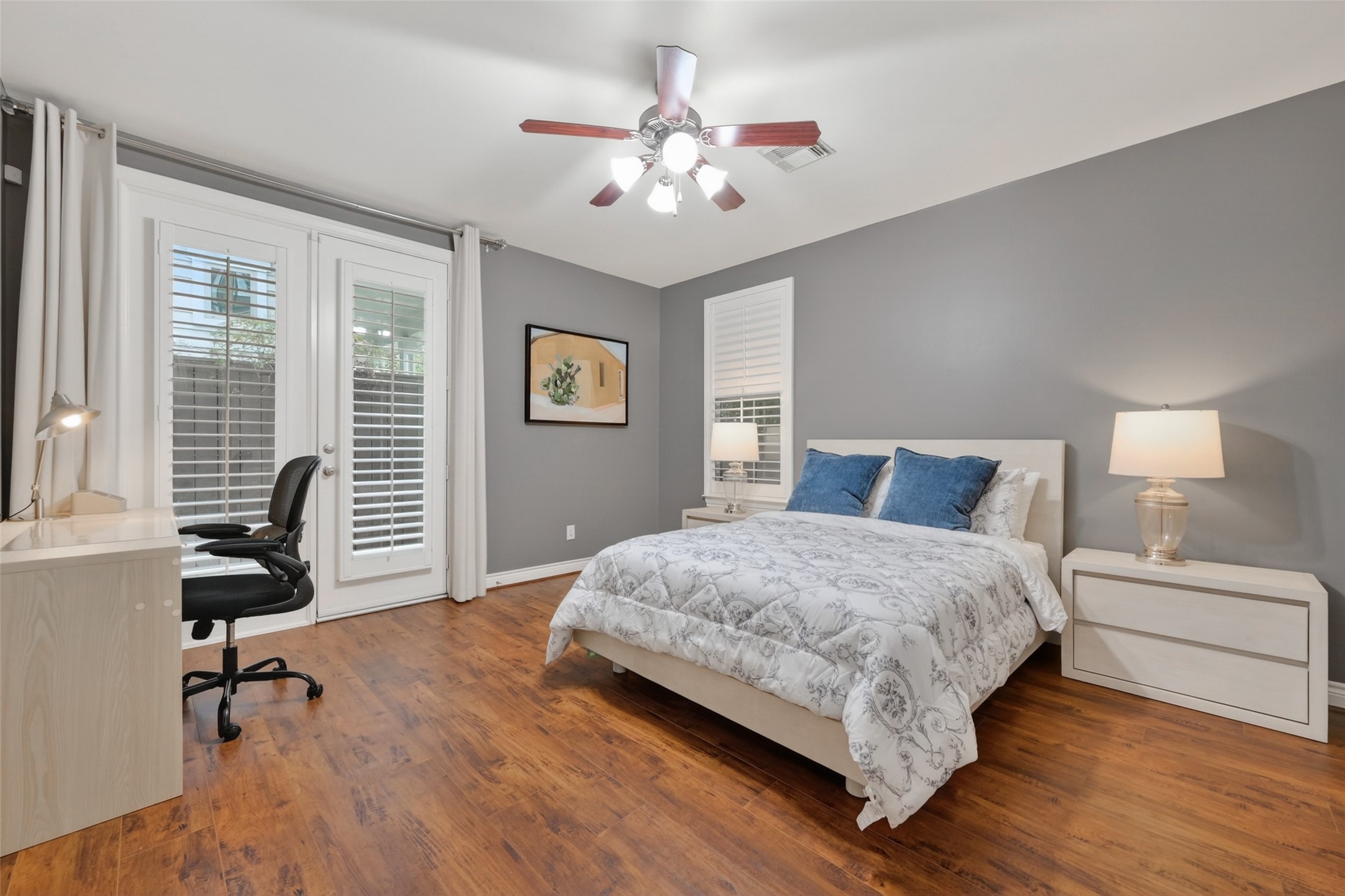 620 Rutland Street, Unit A Houston, TX 77007 - Photo 8 of 46 This spacious bedroom features warm wood flooring, a cozy bed with blue accent pillows, and modern gray walls. It includes a desk with a chair, stylish lamps, and ample natural light through French doors with shutters, creating a comfortable and inviting atmosphere.