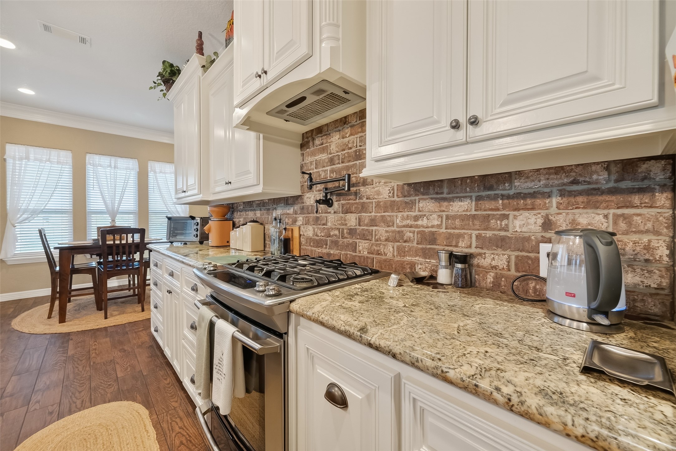 912 County Road 340 Angleton, TX 77515 - Photo 16 of 50 This kitchen features elegant white cabinetry, a rustic brick backsplash, and granite countertops. It includes a gas stove and stainless steel appliances, with a cozy dining area bathed in natural light from multiple windows, all set on warm wood flooring.