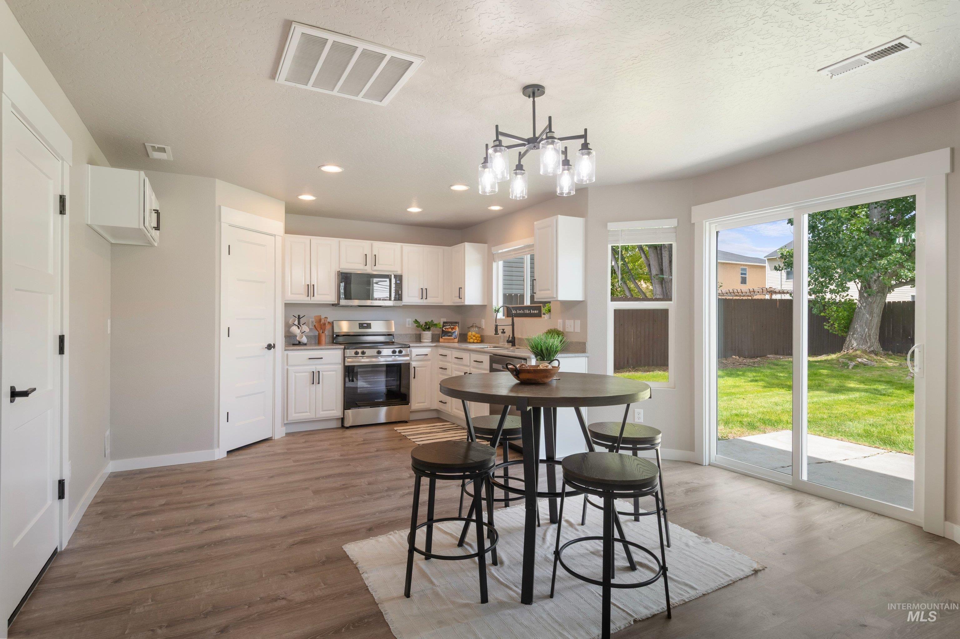 16759 Abram Avenue Caldwell, ID 83607 - Photo 11 of 33 Open-concept kitchen and dining area featuring wood-finish flooring, white cabinetry with stainless steel appliances, a contemporary chandelier, and sliding glass doors opening to a grassy backyard