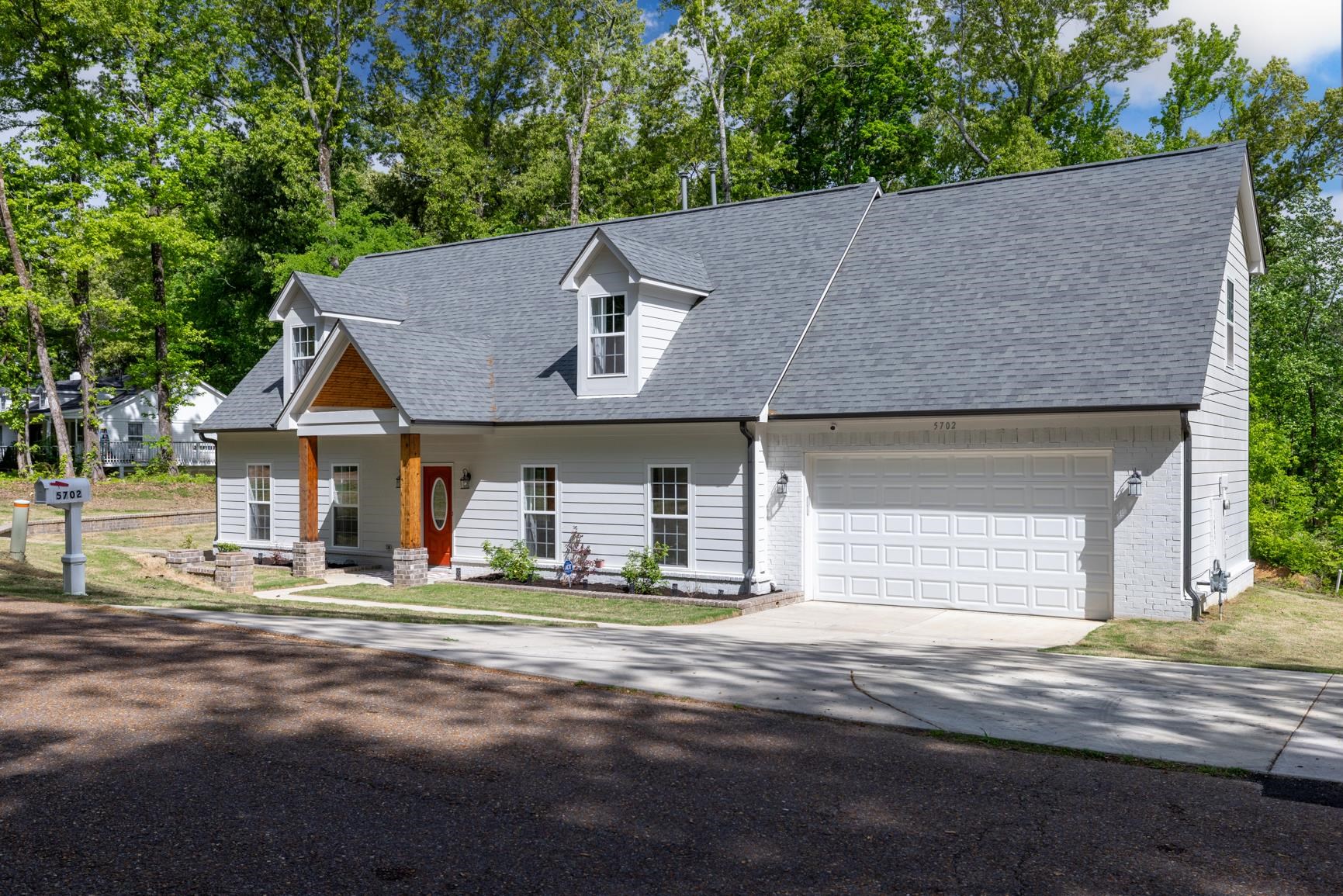 5702 Spring Lake Road Memphis, TN 38135 - Photo 2 of 40 Light gray siding exterior with a dark gray shingle roof, featuring a prominent red front door, dormer windows, a wood-accented porch entry, and an attached two-car garage