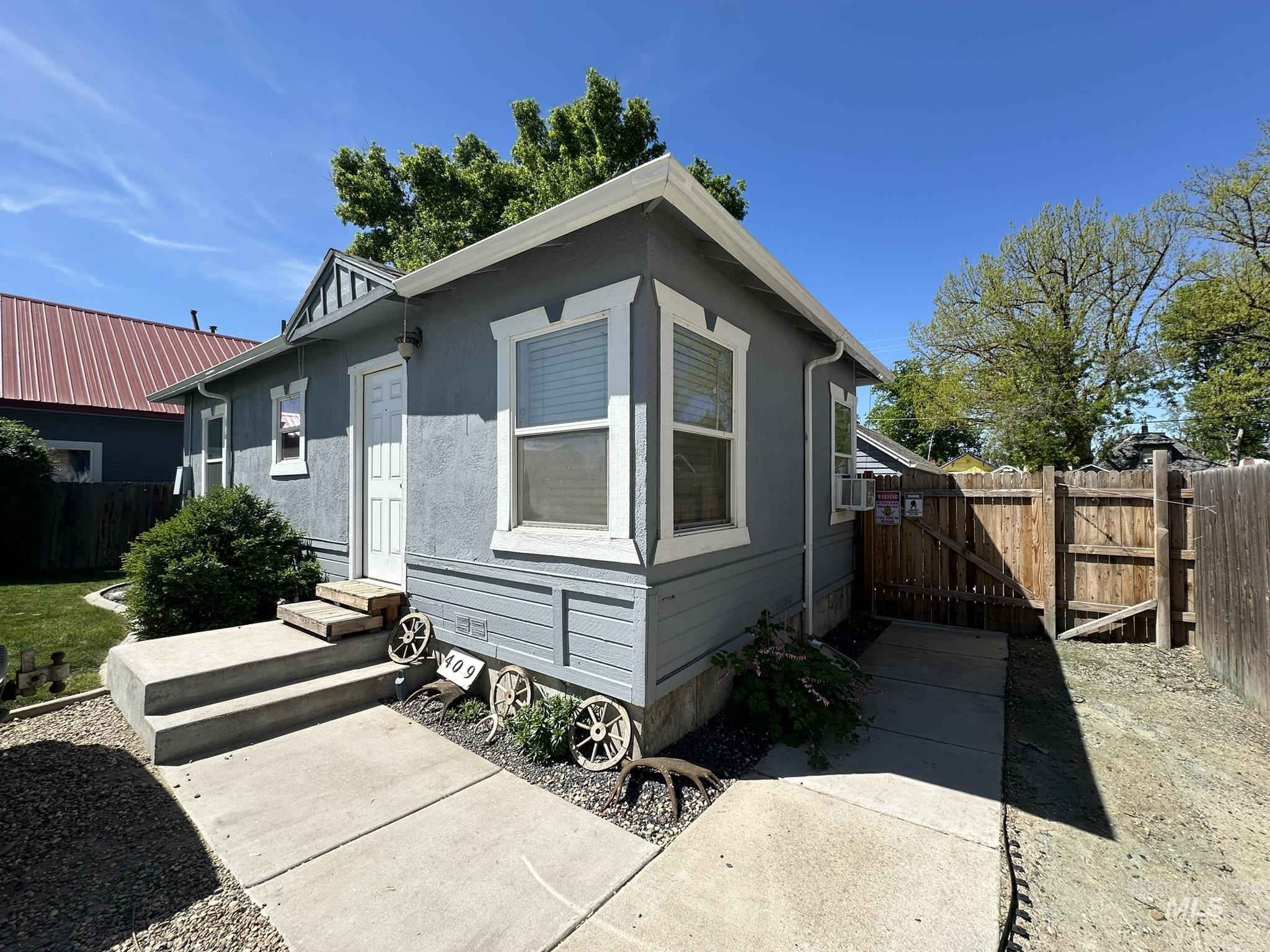 Exterior featuring grey siding, white trim, a concrete walkway, a wood fence, and a mature tree