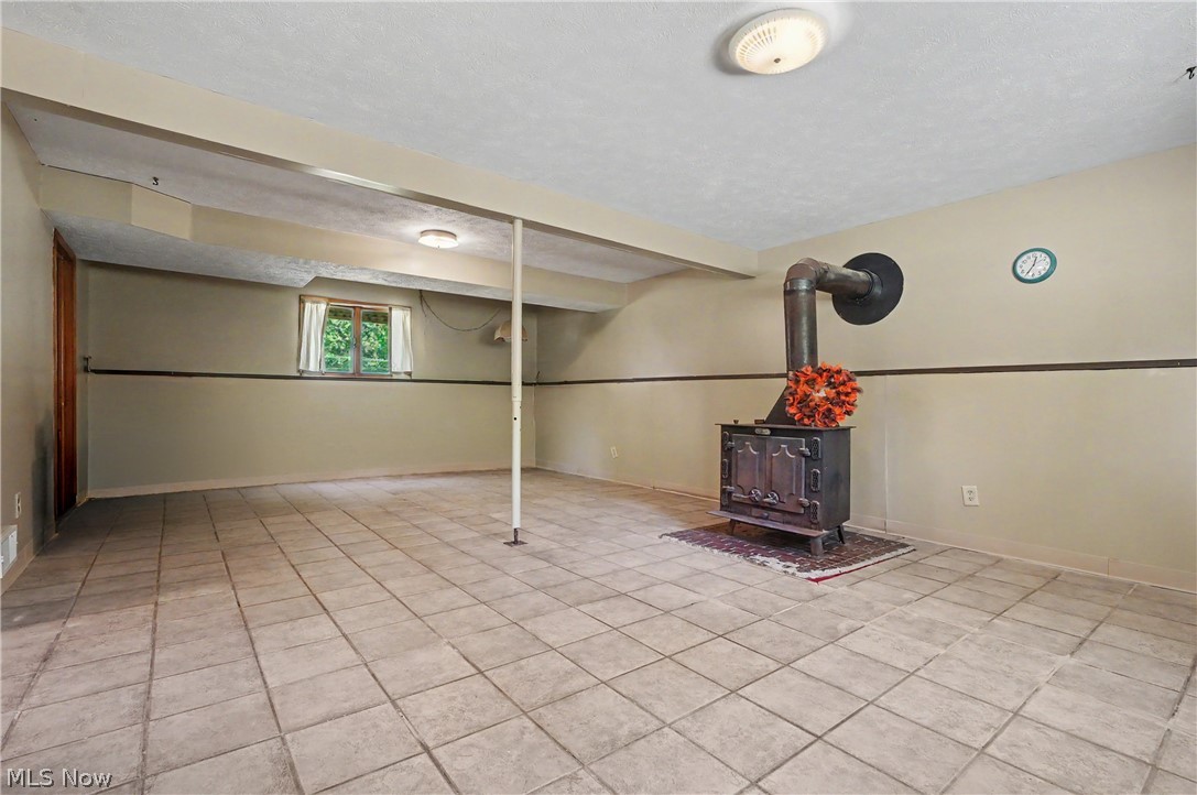 4250 Herr Fieldhouse Road Southington, OH 44470 - Photo 21 of 50 Finished basement space featuring a prominent wood-burning stove with a metal chimney system, large format tile flooring, a window providing natural light, a wood panel door, and overhead lighting fixtures