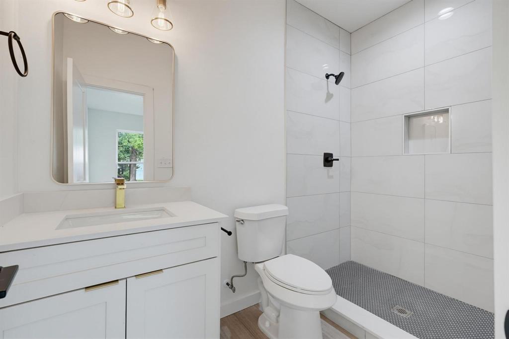 510 South O Connor Road Irving, TX 75060 - Photo 8 of 22 Bathroom featuring a single vanity with a white countertop and gold-toned faucet, a rounded rectangular mirror, and a tiled shower with a built-in niche and dark hardware