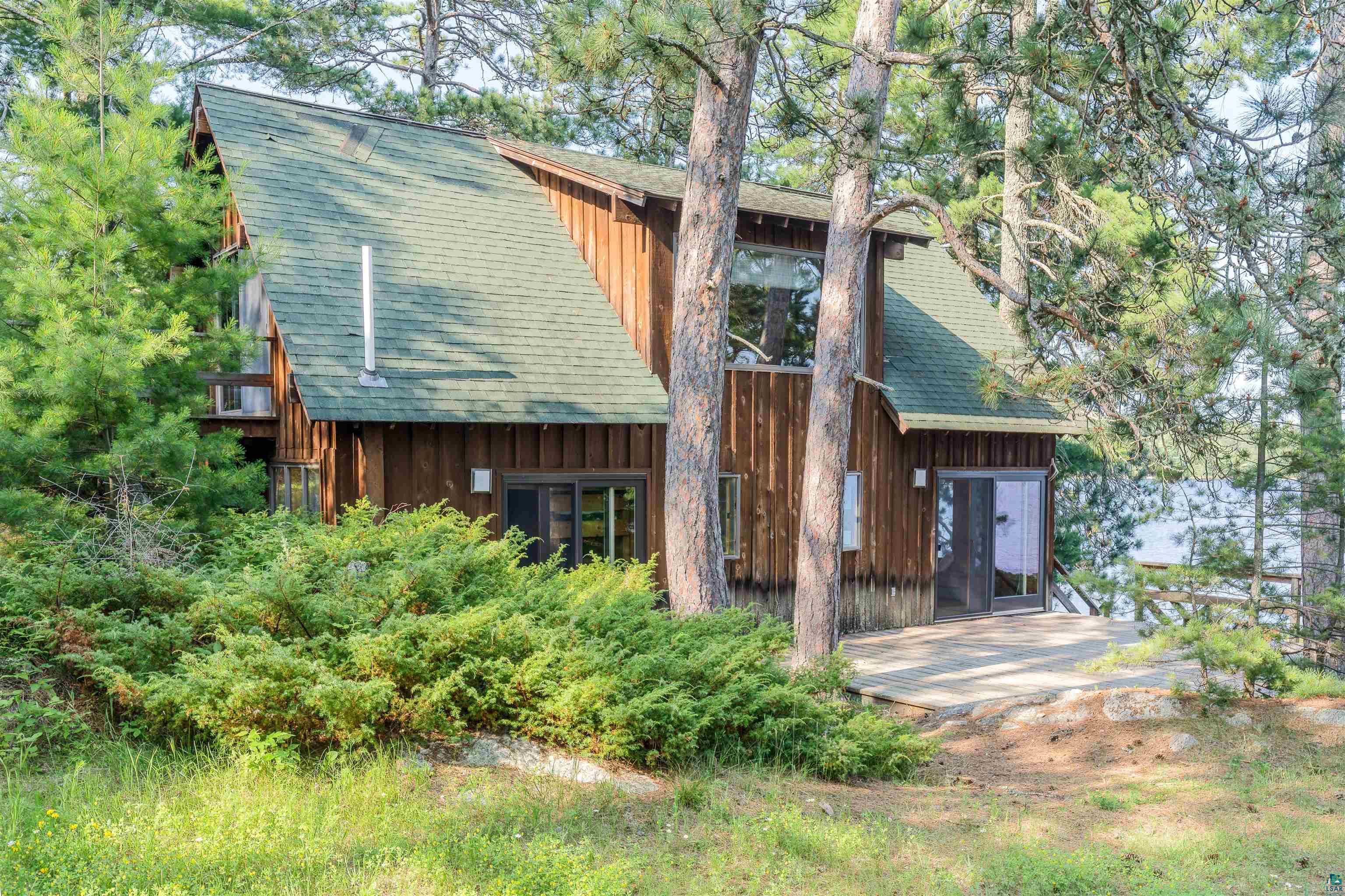 2763 Van Vac Road Ely, MN 55731 - Photo 36 of 37 Wood-sided cabin featuring a steep-pitched green shingled roof, a large picture window, and a ground-level patio with an open sliding glass door