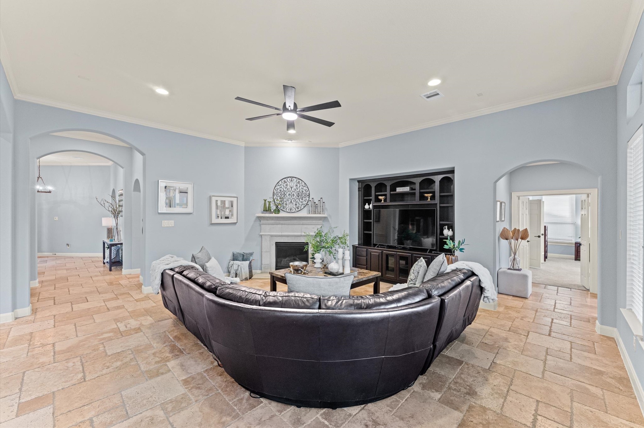 70 Valera Ridge Drive Spring, TX 77389 - Photo 35 of 37 This spacious living room features a stylish curved leather sectional, a modern ceiling fan, and a built-in entertainment center. The light blue walls and tiled flooring create a welcoming atmosphere, complemented by a cozy fireplace. Arched doorways lead to adjacent rooms, enhancing the open-concept feel.