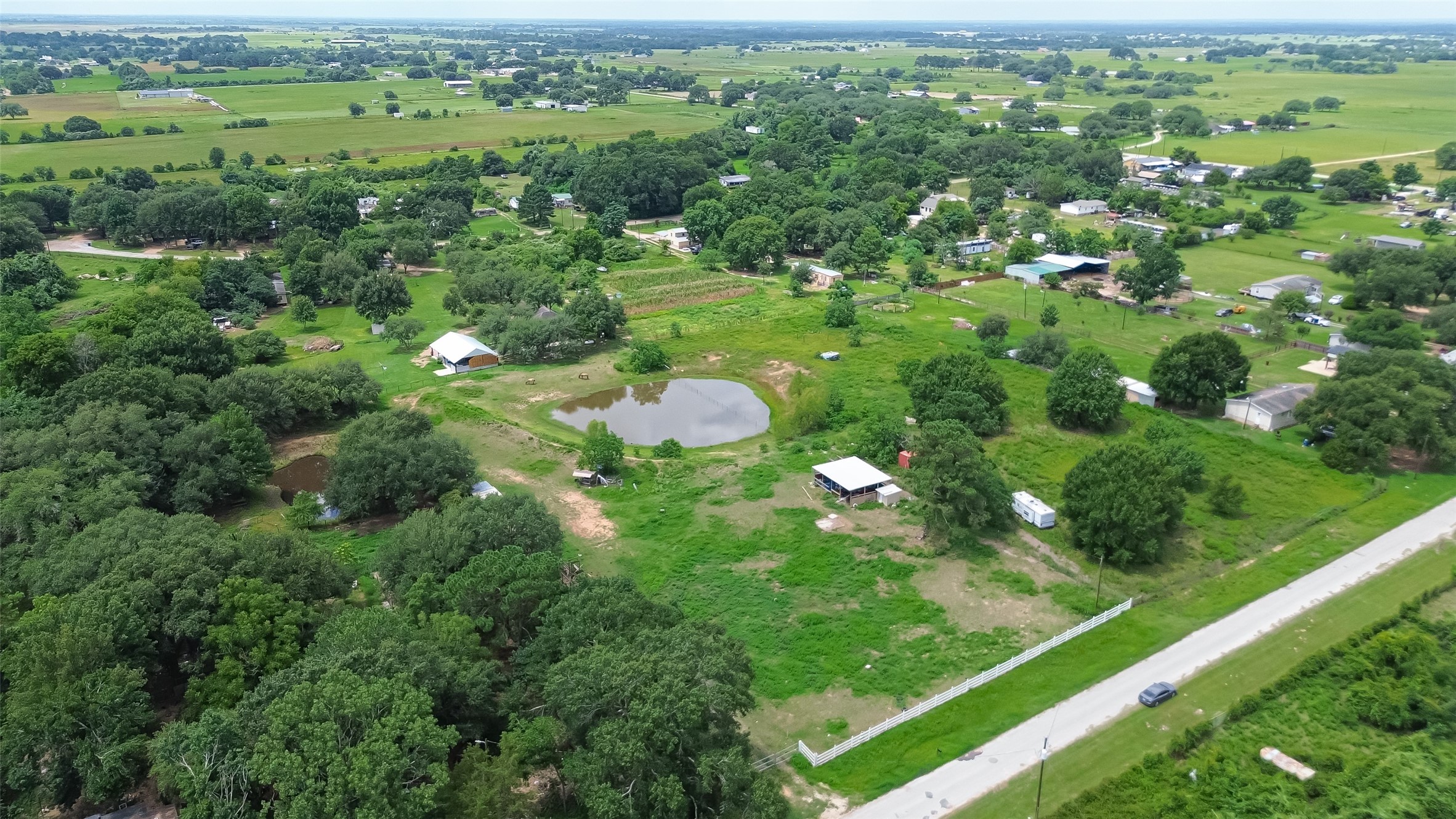 35535 Windmill Road Hempstead, TX 77445 - Photo 26 of 41 This aerial photo showcases a spacious rural property with lush greenery, a central pond, and a few structures. The surrounding area is dotted with trees and open fields, providing a peaceful and private setting. The property is accessible via a nearby road, offering a blend of seclusion and convenience.