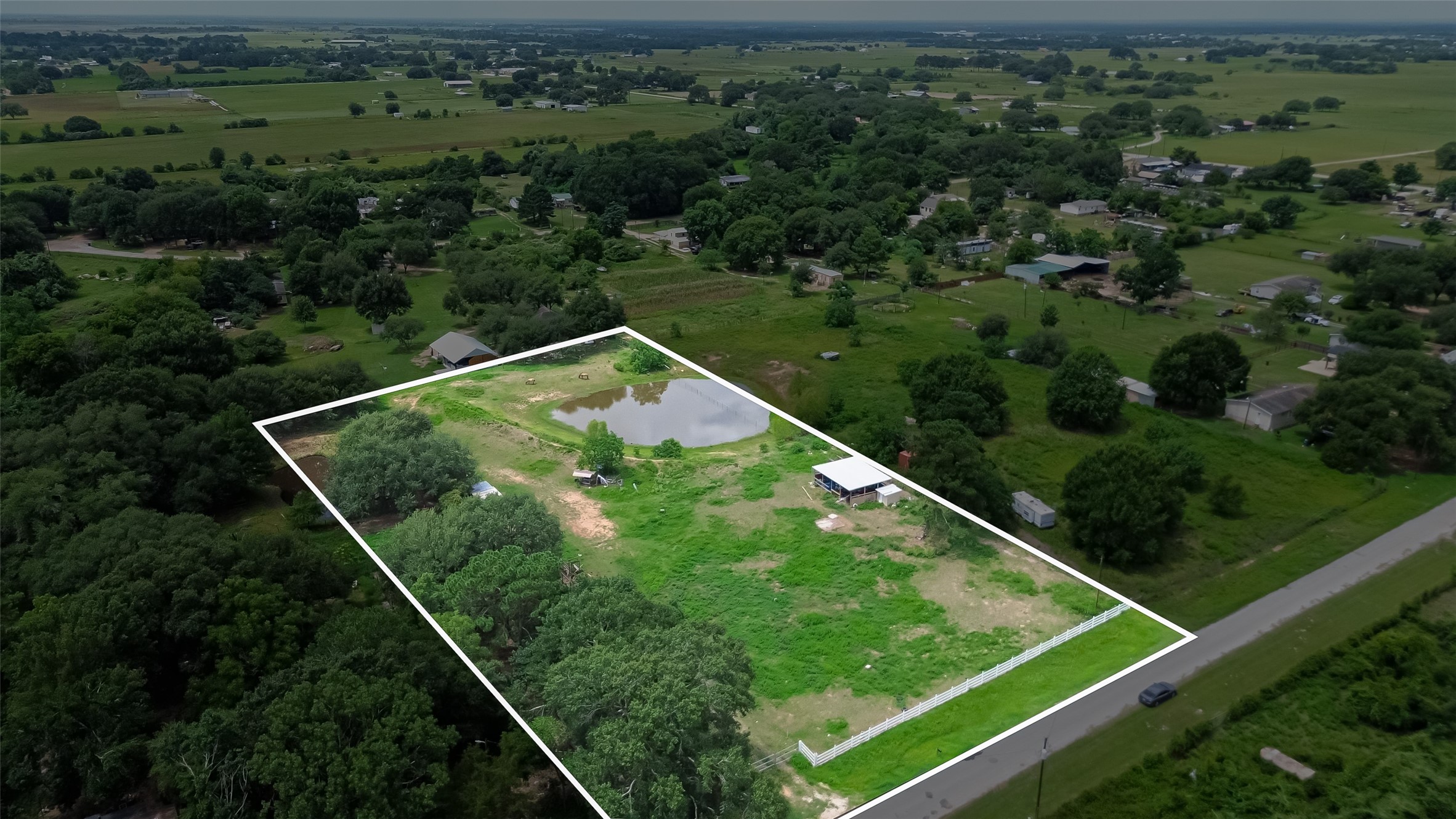 35535 Windmill Road Hempstead, TX 77445 - Photo 8 of 41 This aerial photo showcases a spacious rural property outlined in white, featuring a large open field with scattered trees, a small pond, and a structure. It’s set in a tranquil area with expansive views of surrounding greenery and distant homes, perfect for those seeking privacy and nature.