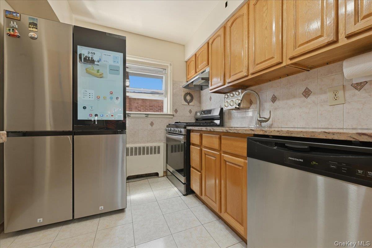 79 Wellington Road Elmont, NY 11003 - Photo 11 of 33 Kitchen featuring stainless steel appliances, wood cabinetry, tile backsplash with accent tiles, light-toned countertops, and tile flooring
