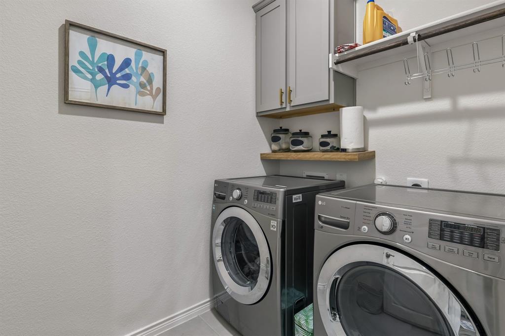 10313 Geode Street Aubrey, TX 76227 - Photo 11 of 13 Dedicated laundry space featuring side-by-side front-loading machines, a wall-mounted wood-finish shelf, and upper cabinetry with gold-tone hardware