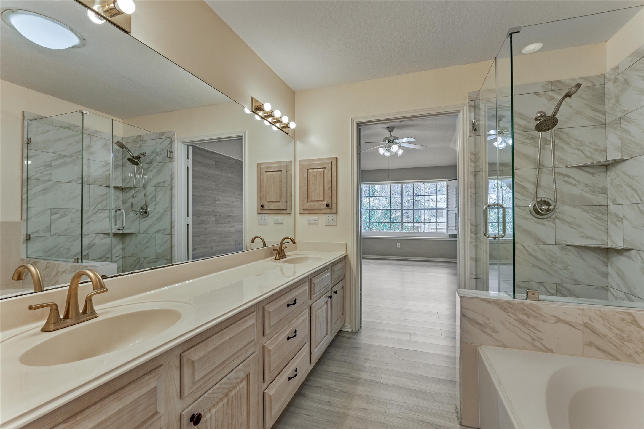 14420 Walters Road, Unit 26 Houston, TX 77014 - Photo 31 of 45 Another view of the primary bathroom with a spacious double vanity and elegant gold fixtures. It features a glass-enclosed shower with marble tiles and a large mirror above the sinks. The room opens to a bright primary bedroom with a ceiling fan and large windows. The overall design is sleek and contemporary.