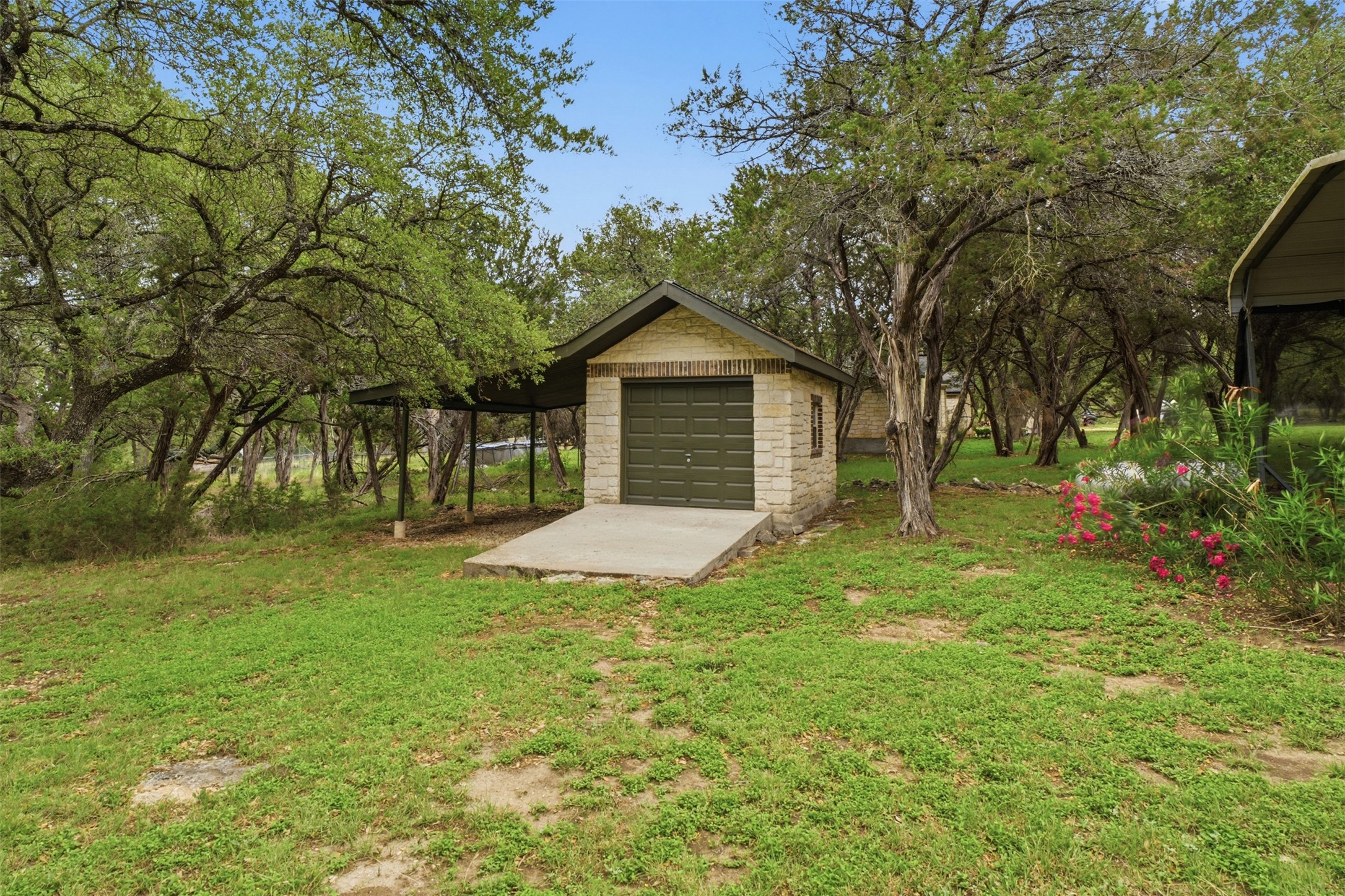 1301 County Road 262 Georgetown, TX 78633 - Photo 33 of 39 Detached stone-clad garage with an olive green garage door and a covered carport, surrounded by mature trees and natural landscaping