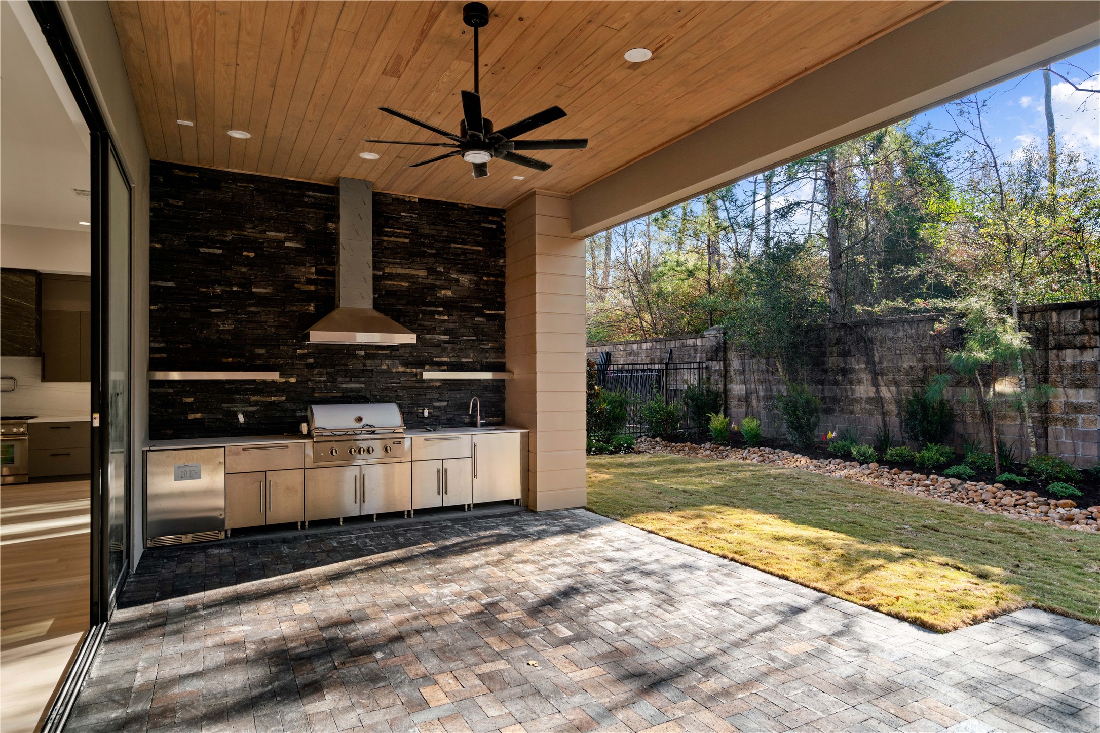 47 Pronghorn Place Spring, TX 77389 - Photo 40 of 44 This photo showcases a modern outdoor kitchen and patio area. The space features a built-in grill, stainless steel cabinetry, and a sleek black backsplash. The ceiling is wood-paneled with recessed lighting and a ceiling fan. It opens up to a landscaped backyard with a stone border, creating an inviting space for outdoor entertaining.
