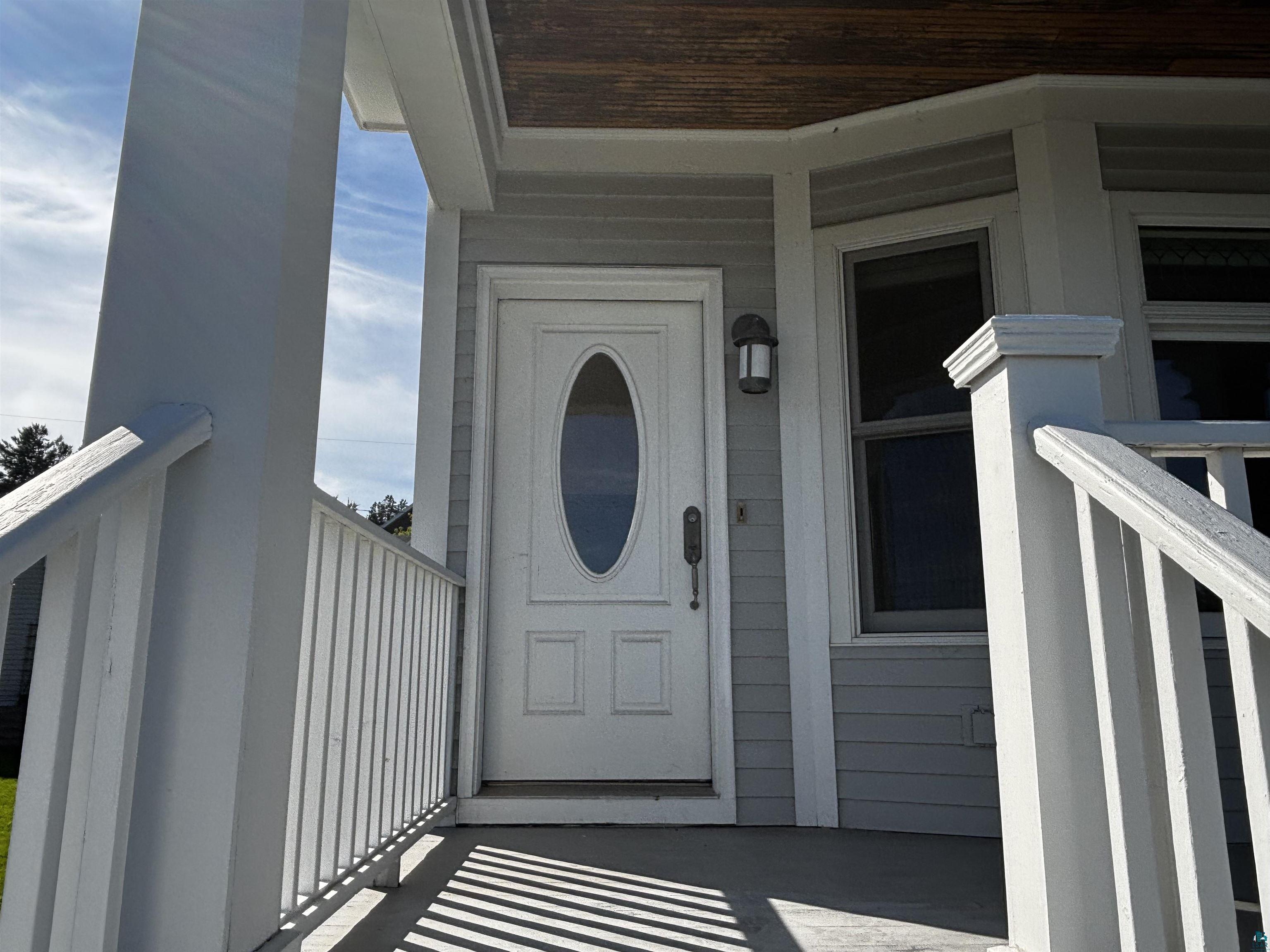 13 West 8th Street Duluth, MN 55806 - Photo 49 of 57 Covered front porch with white railing, featuring a panel door with an oval-shaped glass insert and an exterior wall sconce