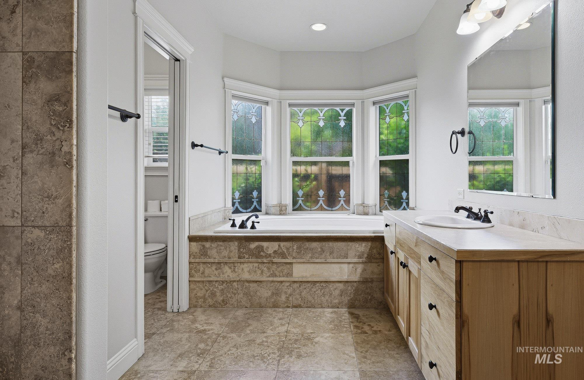 2172 West Cedar Grove Street Meridian, ID 83646 - Photo 15 of 49 Expansive bathroom featuring a built-in soaking tub with a tiled surround, three bay windows with decorative glass accents, a large wood-finish vanity with an integrated sink, and stone-finish floor tiling