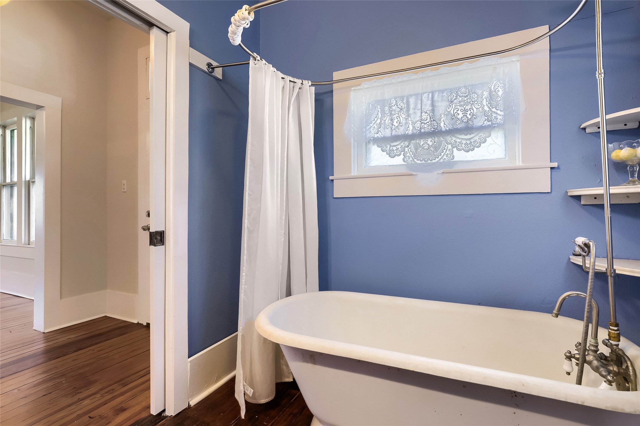 208 North Ave B Elgin, TX 78621 - Photo 25 of 29 Bathroom 2 features a charming clawfoot tub with a shower curtain, set against blue walls. It has a small window with lace curtains and wooden floors, creating a cozy, classic feel.