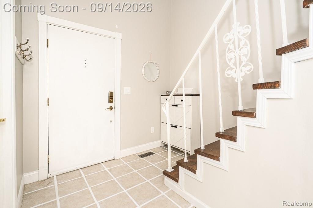 Entry foyer with square tile flooring, a white entry door with brass hardware, and a staircase featuring wood treads and decorative metal balusters