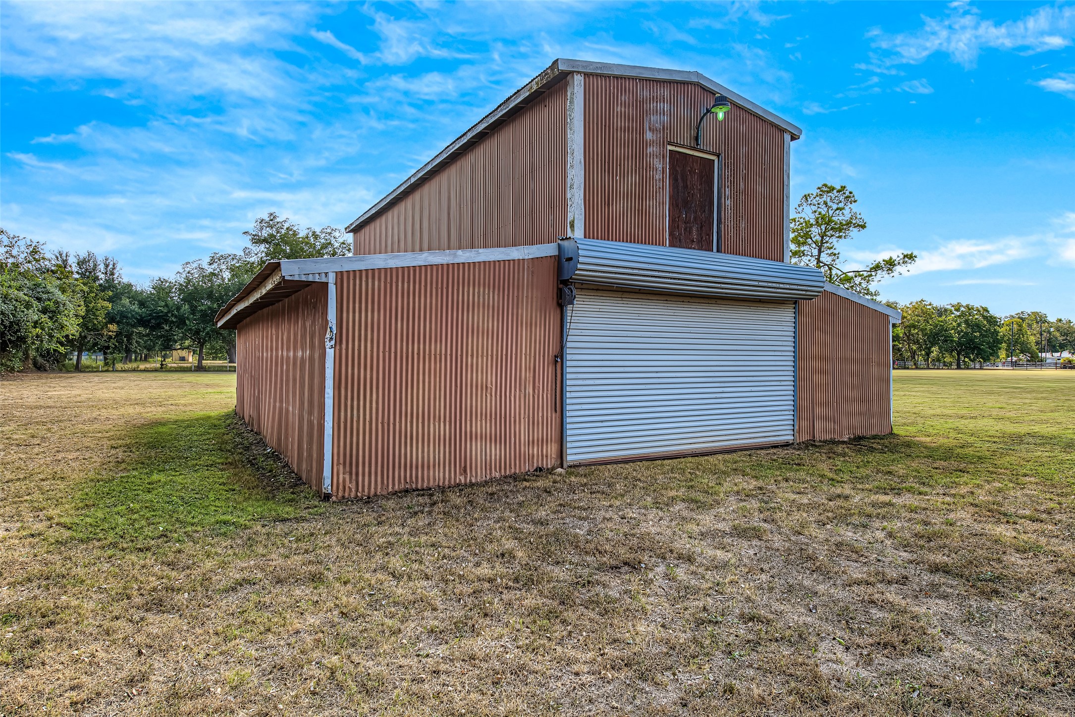 1345 Peach Ridge Road Brookshire, TX 77423 - Photo 42 of 50 A standout asset for country living, this substantial freestanding metal barn/workshop features durable corrugated metal siding, a wide metal roll-up door for easy vehicle or equipment entry, and a upper loft level complete with top-door access and exterior gooseneck lighting. Enclosed by lean-to side wings for additional covered workspace or storage, this versatile structure is ideal for housing tractor equipment, workshop tools, boats, RV storage, or agricultural supplies.