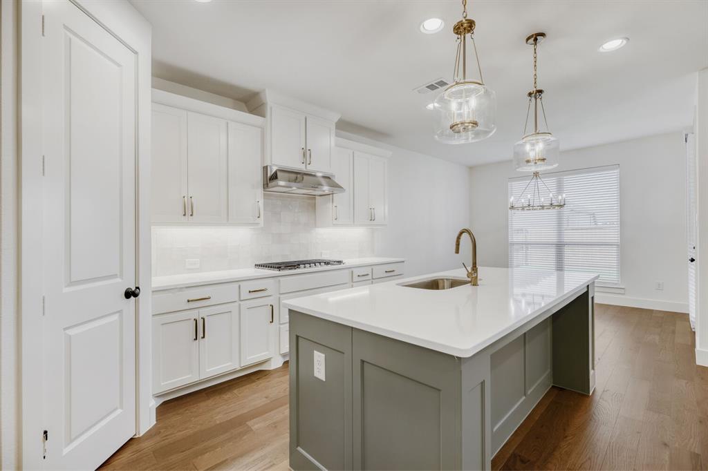 4314 Haley Way Oak Point, TX 75068 - Photo 9 of 28 Modern kitchen featuring a large island with sink and bronze-finish faucet, white shaker cabinetry with under-cabinet lighting, stainless steel range hood, and wood-finish flooring
