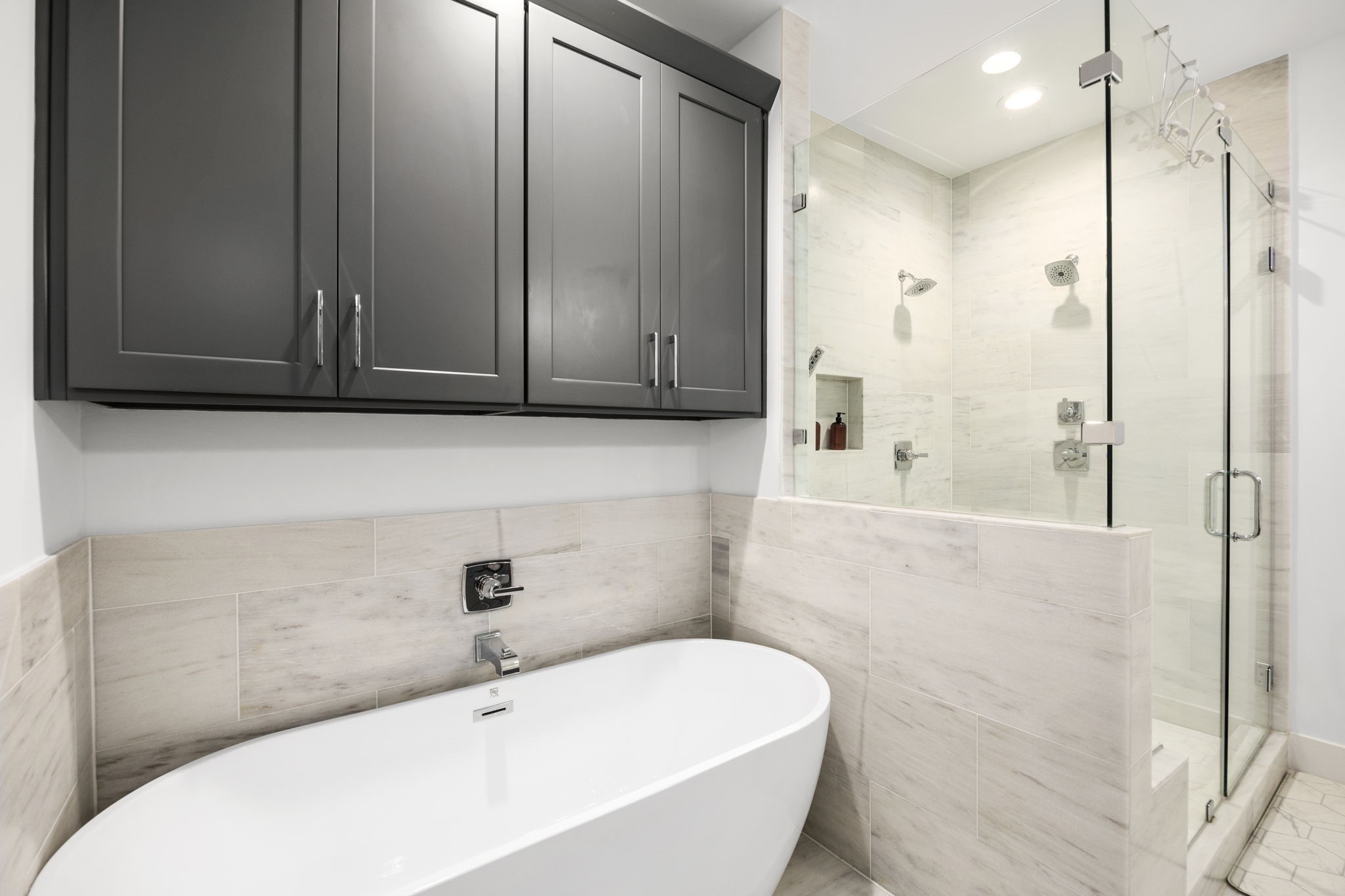 2411 Hazard Street Houston, TX 77019 - Photo 32 of 46 This close-up view of the primary bathroom highlights the freestanding soaking tub paired with sleek, modern fixtures. Above the tub, dark cabinetry provides extra storage space, complementing the neutral-toned tile work. Adjacent to the tub, the spacious glass-enclosed shower features dual shower heads and a built-in niche for added functionality. The clean lines and sophisticated design create a relaxing, spa-like atmosphere.