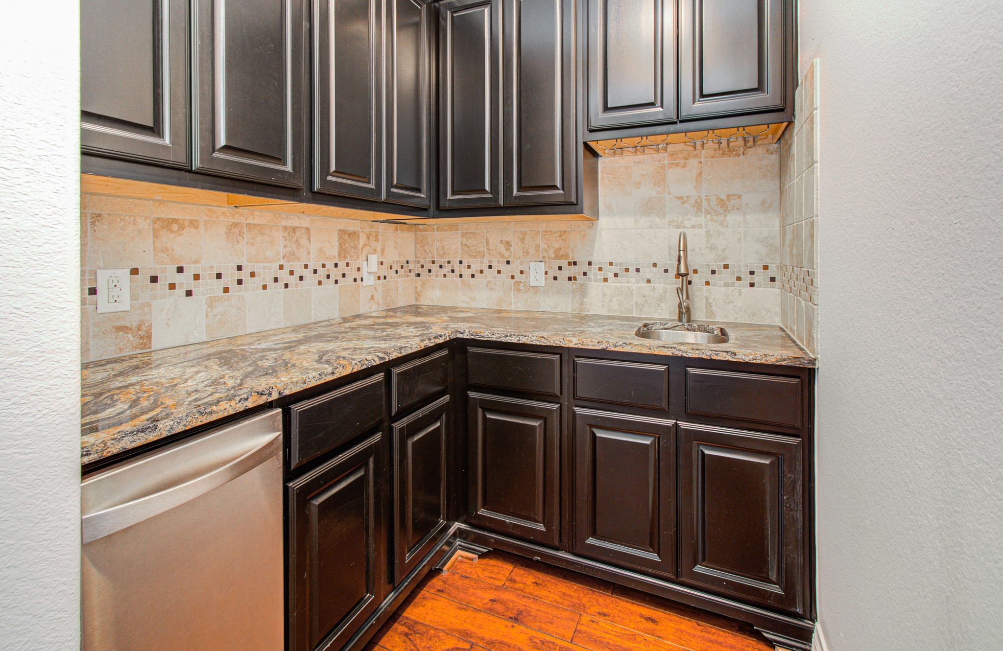 6621 North Main Street Houston, TX 77009 - Photo 5 of 41 This photo showcases a compact, stylish bar area with dark wooden cabinets, granite countertops, and a tile backsplash. There's a stainless steel dishwasher and a small sink, all complemented by warm wood flooring.