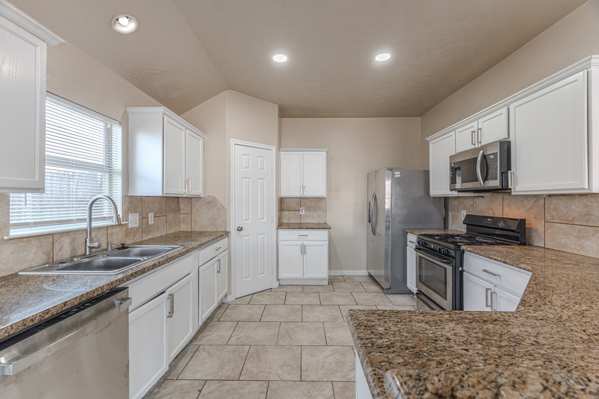 3619 Fiorella Way Humble, TX 77338 - Photo 5 of 16 This kitchen features modern appliances, white cabinetry, and granite countertops. It has a spacious layout with a double sink, a gas range, and ample natural light from a large window. The tile flooring complements the overall clean and bright aesthetic.