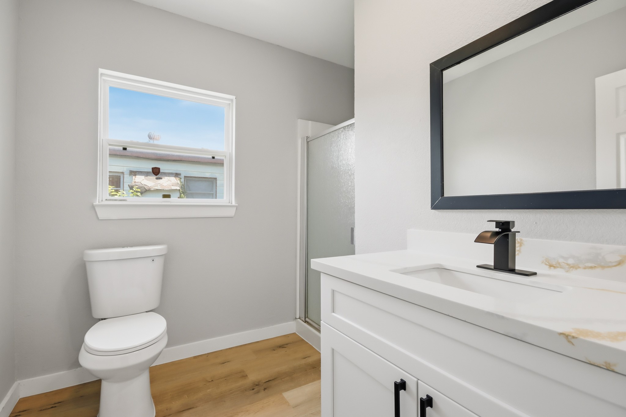 10430 Wiggins Street Houston, TX 77029 - Photo 21 of 27 Secondary bathroom features a modern design with brand new wood-look laminate floor. It includes a sleek vanity with a black faucet, a large mirror, and a window providing natural light. The shower area is enclosed, creating a clean and functional space.