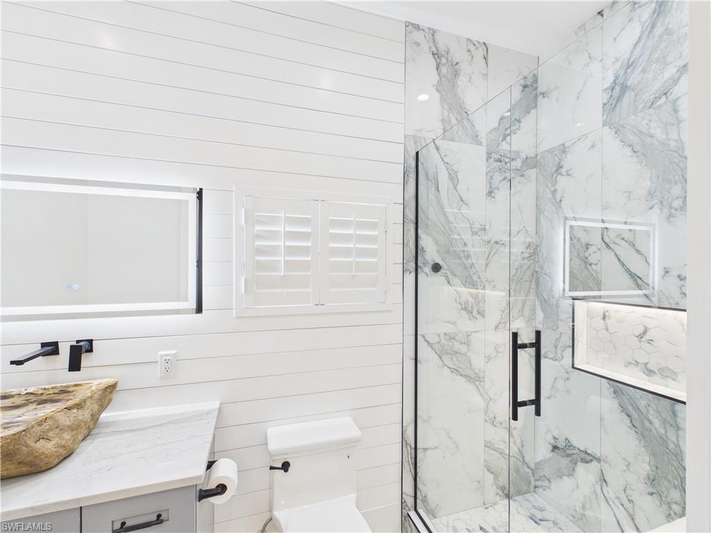3650 Sunset Boulevard Naples, FL 34112 - Photo 21 of 26 Bathroom featuring shiplap wall paneling, a vessel sink with a wall-mounted faucet, a vanity with a stone countertop, a glass-enclosed shower with marbled tile, and an illuminated shower niche