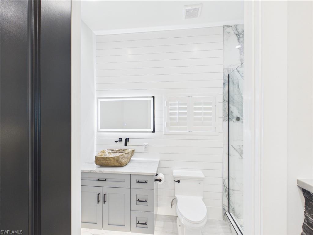 3650 Sunset Boulevard Naples, FL 34112 - Photo 22 of 26 Bathroom featuring a unique stone vessel sink, gray vanity with dark hardware, illuminated mirror, and a glass-enclosed shower with marble-look wall tile