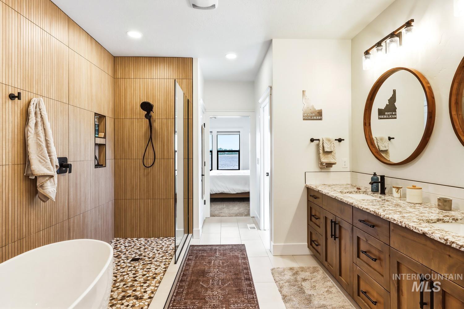 18658 Tranquil Place Caldwell, ID 83607 - Photo 26 of 44 Bathroom featuring a freestanding soaking tub, a walk-in shower with pebble flooring and wood-finish wall tile, and a dual vanity with granite countertops and round wooden mirrors