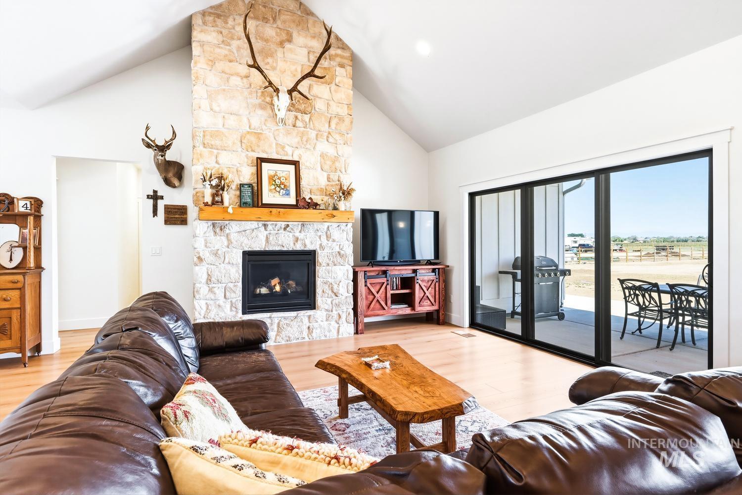 18658 Tranquil Place Caldwell, ID 83607 - Photo 6 of 44 Vaulted living area featuring a stone-clad fireplace with a natural wood mantel, light wood-finish flooring, and expansive multi-panel sliding glass doors opening to an outdoor patio with distant fields