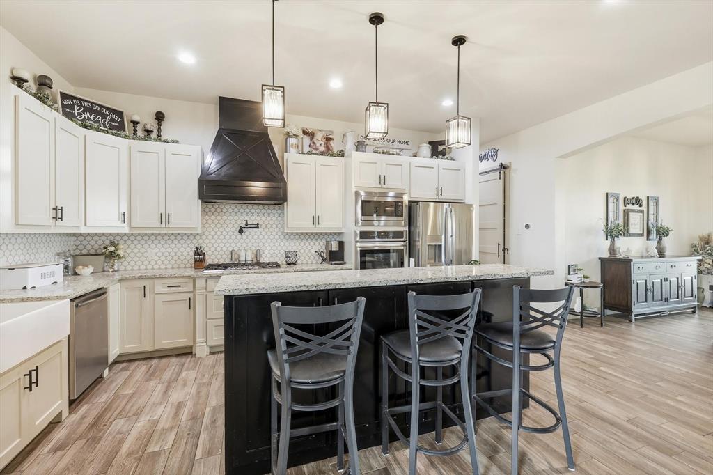 6020 Honeysuckle Road Midlothian, TX 76065 - Photo 10 of 39 Kitchen featuring a large island with counter seating, a white apron-front sink, stainless steel appliances, a black range hood, and wood-finish flooring