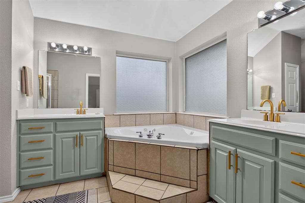 100 Radecke Road Krum, TX 76249 - Photo 25 of 29 Corner soaking tub with tiled surround, flanked by two vanities featuring light green cabinetry, white countertops, and gold-toned fixtures