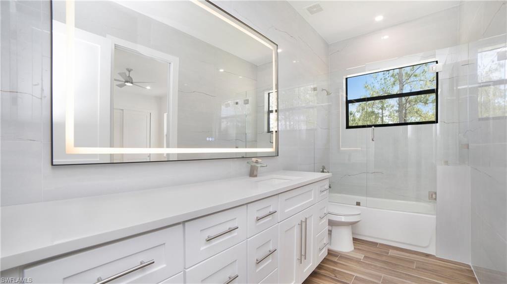 2343 Oil Well Road Naples, FL 34120 - Photo 21 of 46 Contemporary bathroom featuring a spacious vanity with white cabinetry and a light-toned countertop, an illuminated rectangular mirror, a white bathtub with a glass shower panel, and wood-finish tile flooring