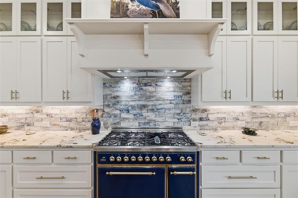 6625 Sandy Beach Road Azle, TX 76020 - Photo 12 of 40 Kitchen cooking area features commercial-style gas range with double ovens, custom vent hood surround, textured tile backsplash, extensive cabinetry storage, and stone countertop surfaces extending throughout the surrounding prep areas.