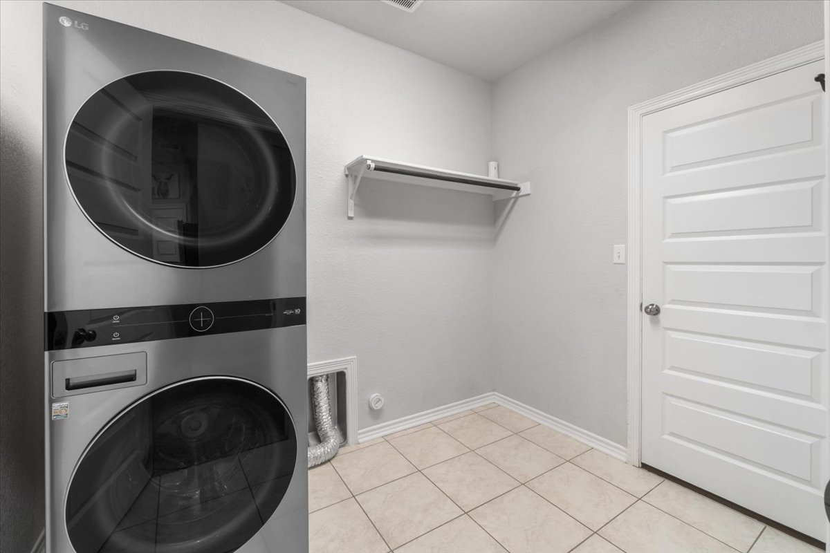 5408 Vanner Path Georgetown, TX 78626 - Photo 29 of 34 Laundry area featuring a stacked washer and dryer unit, neutral ceramic tile flooring, a white paneled interior door, a wall-mounted shelving unit, and light gray wall paint