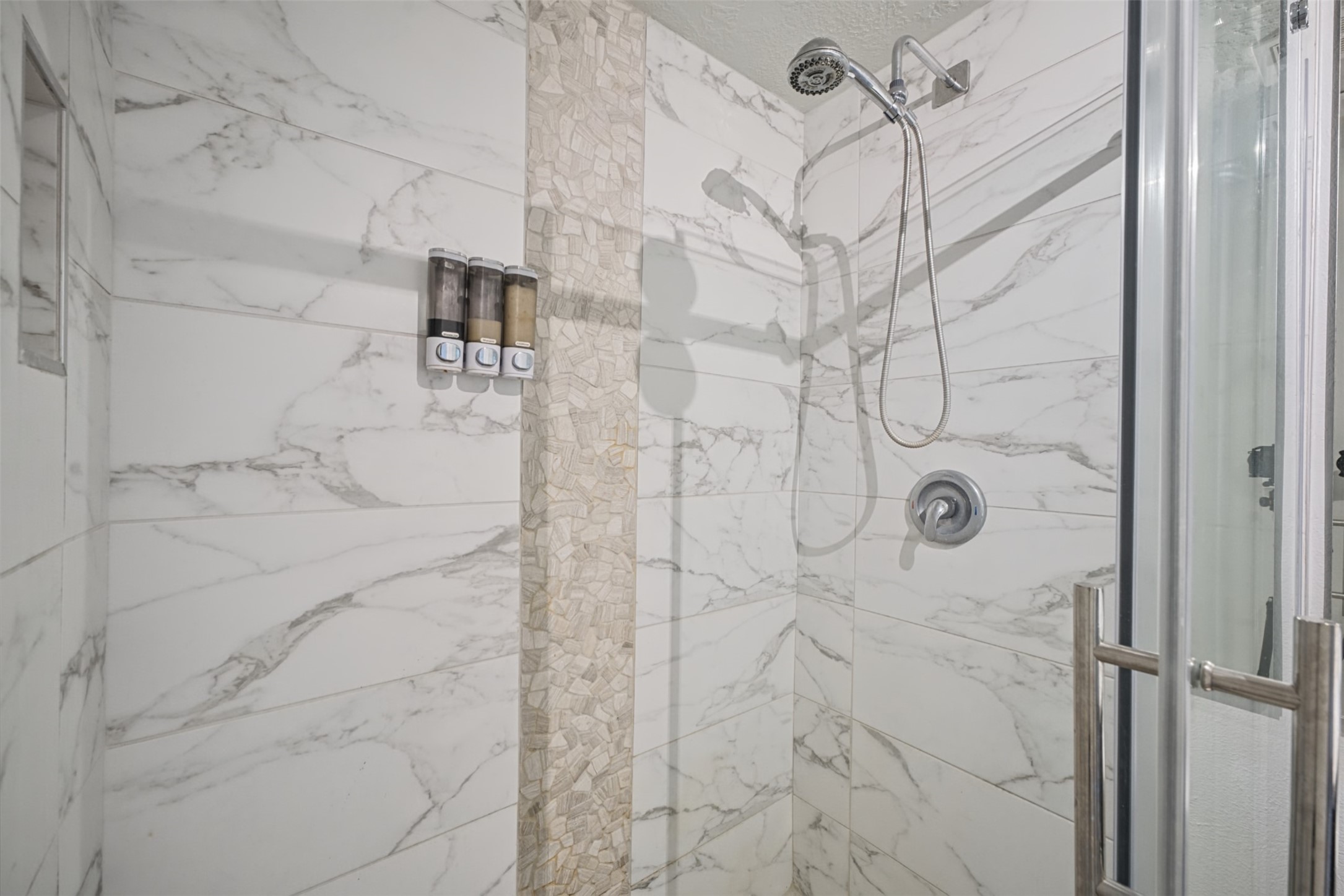 801 River Road, Unit 112F Montgomery, TX 77356 - Photo 17 of 27 Spa-inspired walk-in shower features striking marble-look tile with a decorative stone accent, frameless glass enclosure, and multiple shower heads, including a convenient handheld shower.