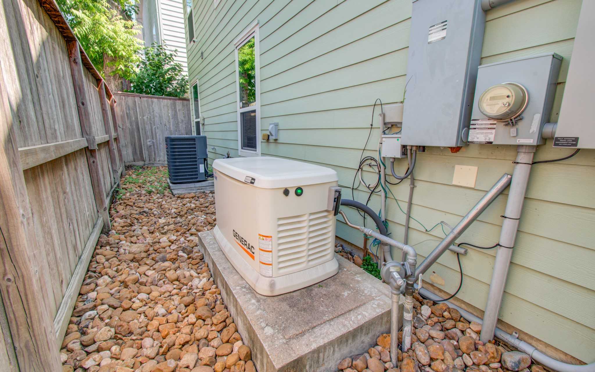 6621 North Main Street Houston, TX 77009 - Photo 30 of 41 This photo shows the side of a house with an installed Generac backup generator, an air conditioning unit, and utility meters. The area is bordered by a wooden fence and features a stone ground covering, providing a neat and functional space for essential home utilities.