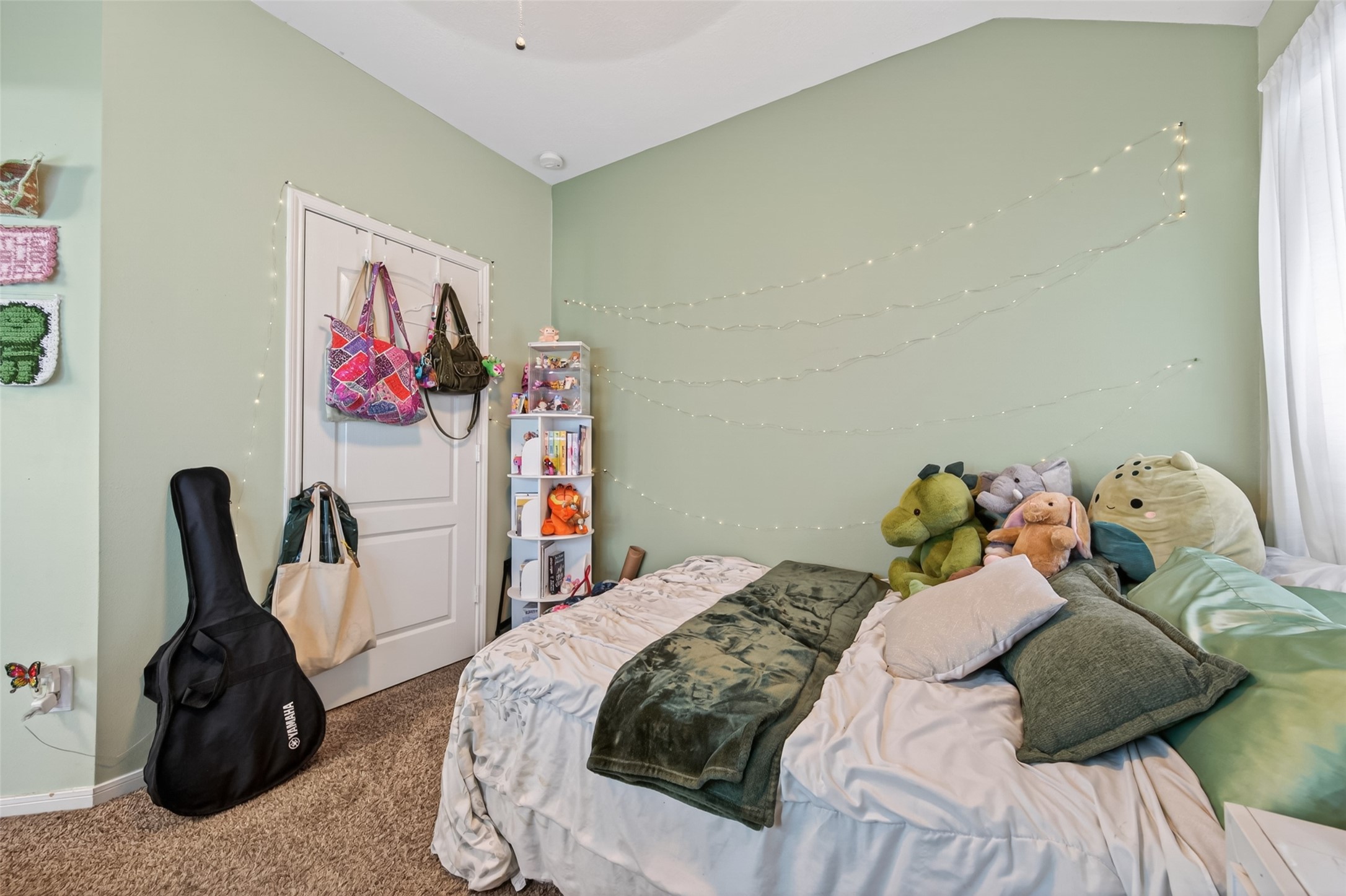 8622 Henrico Lane Rosenberg, TX 77469 - Photo 15 of 20 This cozy bedroom features soft green walls and carpeted flooring. It includes a comfortable bed adorned with plush toys, a small shelf for storage, and decorative string lights. A guitar case and bags hang neatly on the door, adding a personal touch.
