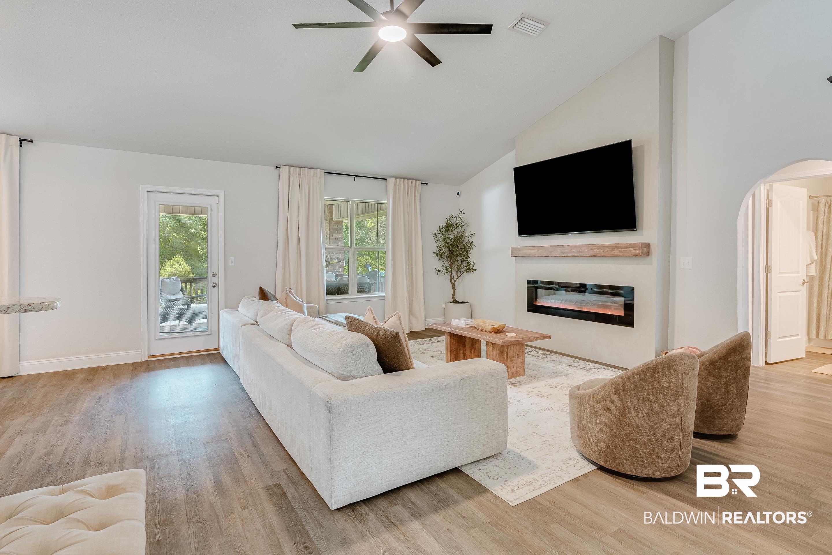 2215 Fernwood Loop West Semmes, AL 36575 - Photo 7 of 40 Living room featuring wood-finish flooring, a wall-mounted electric fireplace with a wood mantle, a ceiling fan, a glass-paned door to the exterior, and a large window with white drapery