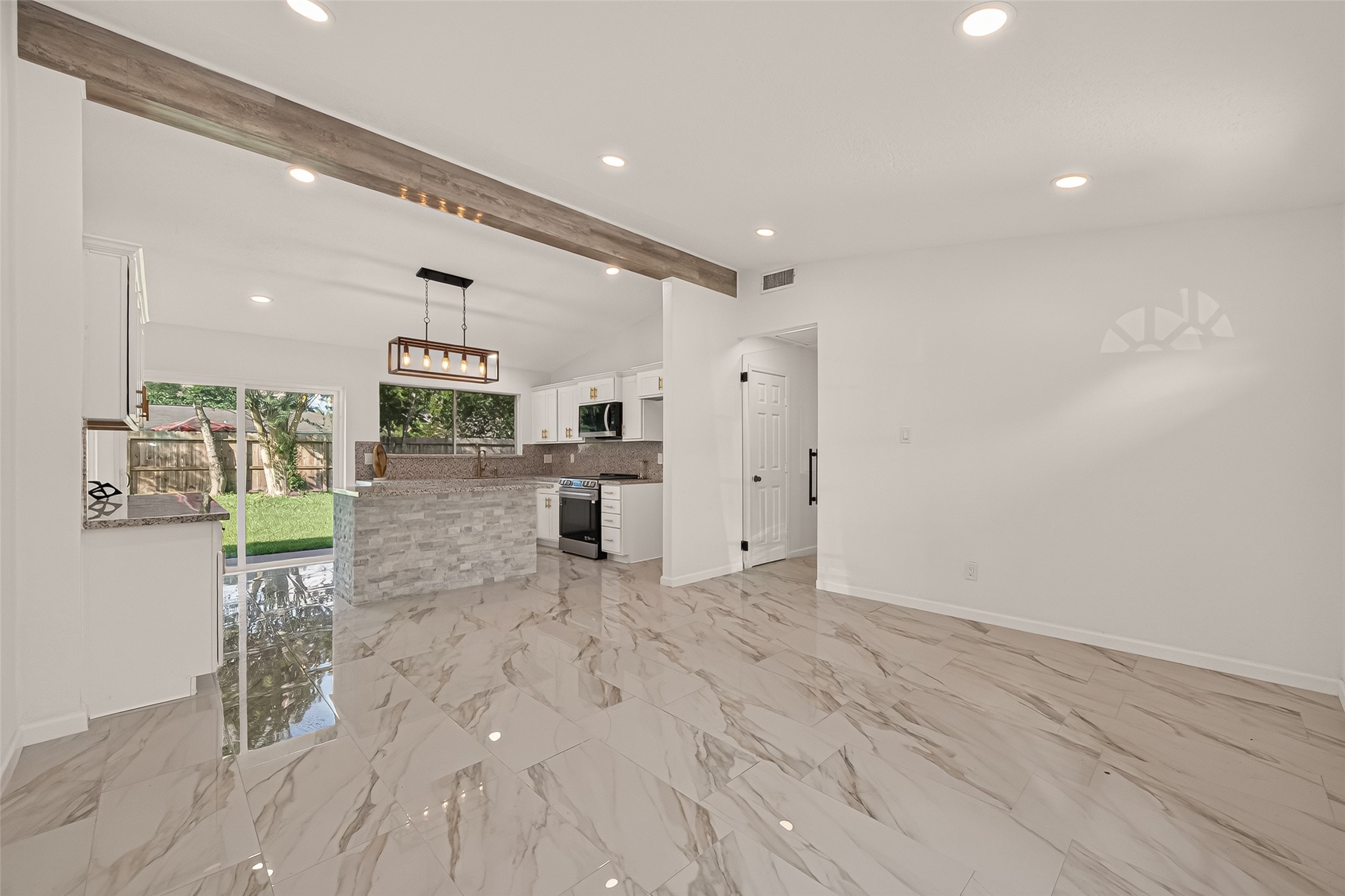 16619 Tibet Road Friendswood, TX 77546 - Photo 5 of 29 This photo showcases a modern open-concept kitchen and living space with elegant marble tile flooring, recessed lighting, and a stylish wood beam accent. The kitchen features a central island with stone detailing, contemporary light fixtures, and large windows offering plenty of natural light and views of the backyard.