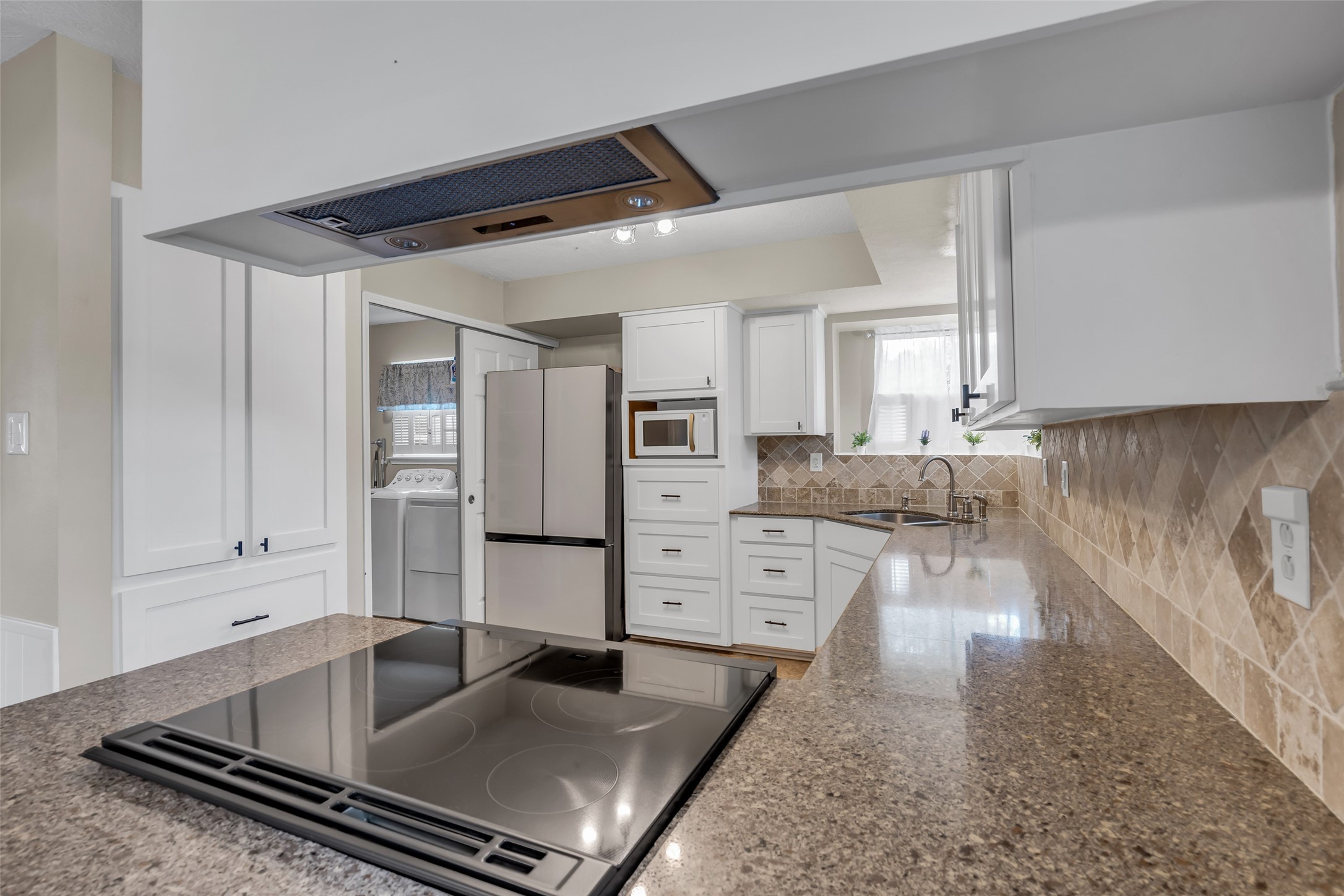 22502 Fields Lane Spring, TX 77389 - Photo 14 of 39 Modern kitchen with granite countertops, a smooth cooktop, and white cabinetry. There's a built-in oven, microwave, and a stainless steel refrigerator. The space is bright with natural light and features a stylish tile backsplash. A laundry area is visible in the adjacent room.