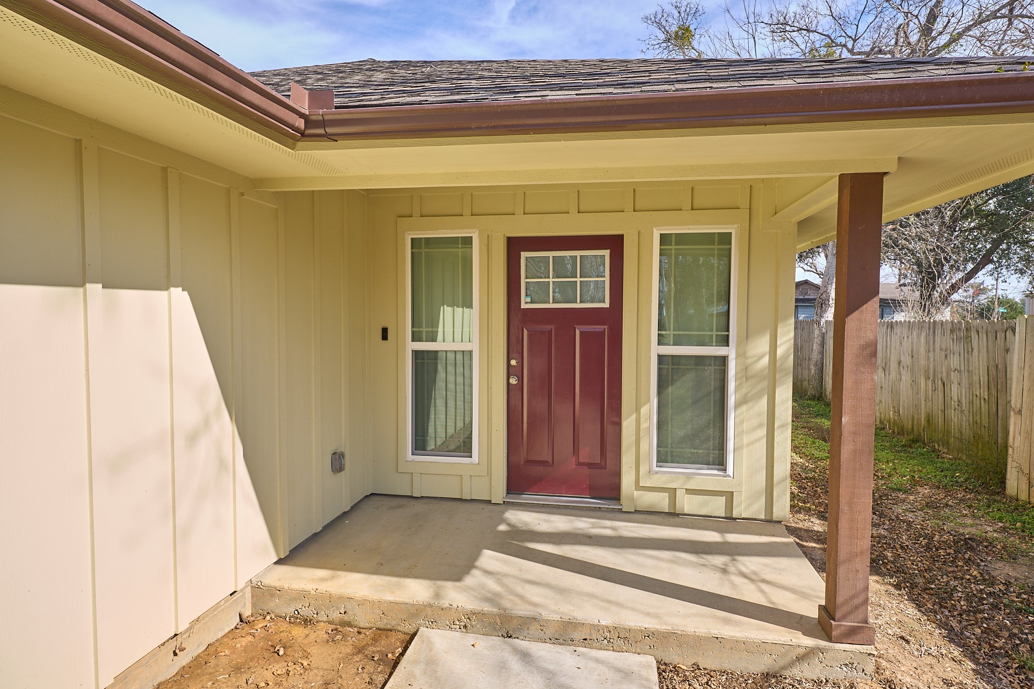 1807 Nuches Lane Bryan, TX 77803 - Photo 4 of 27 This photo shows a welcoming front porch featuring a red door flanked by two large windows. The exterior has a light beige color with brown trim, and there's a small concrete patio area leading up to the entrance. The space is partially covered, providing some shelter.