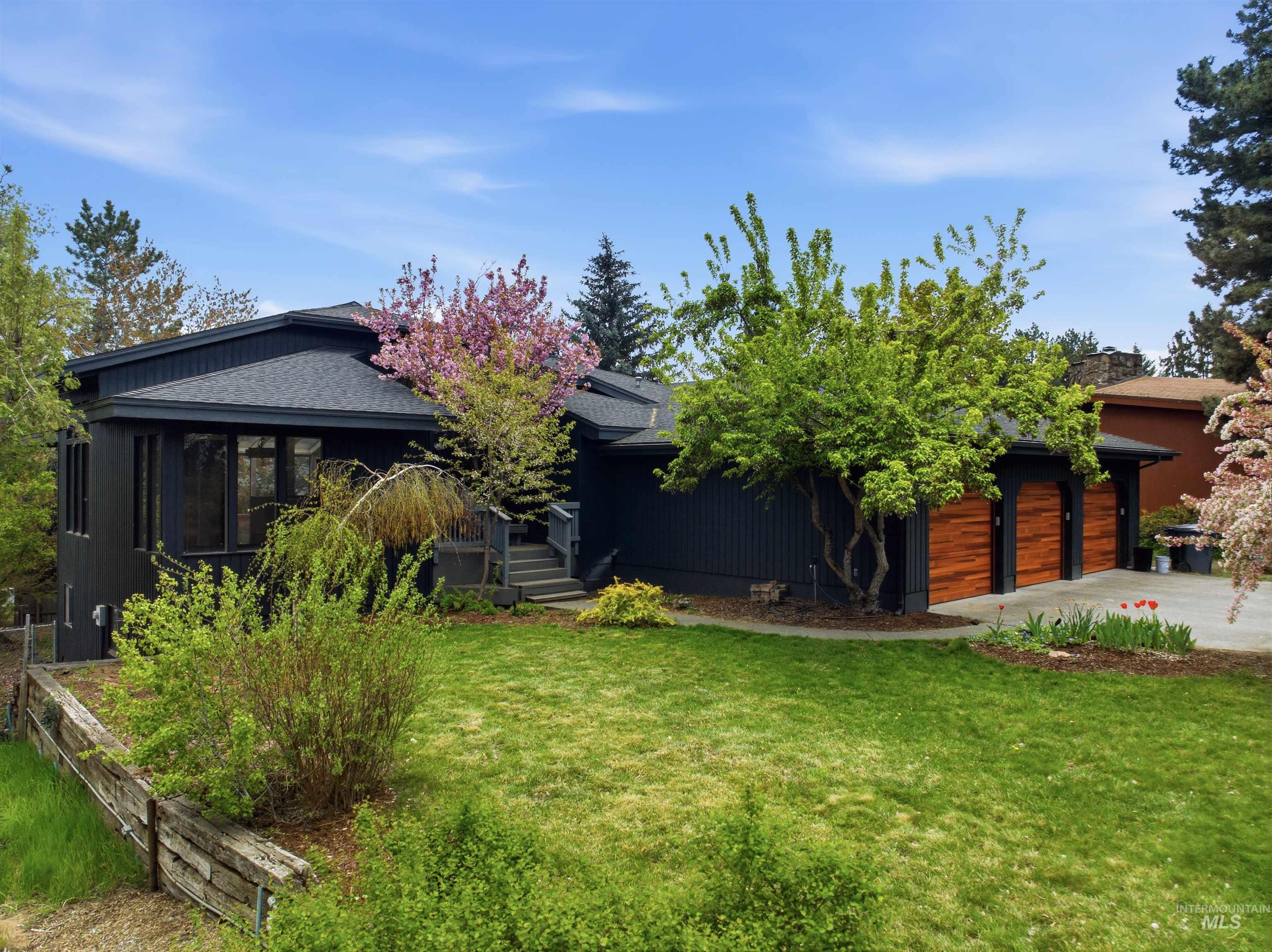 Contemporary dark-paneled exterior featuring a multi-level roofline, a prominent two-car garage with wood-finish doors, a verdant lawn, and established trees