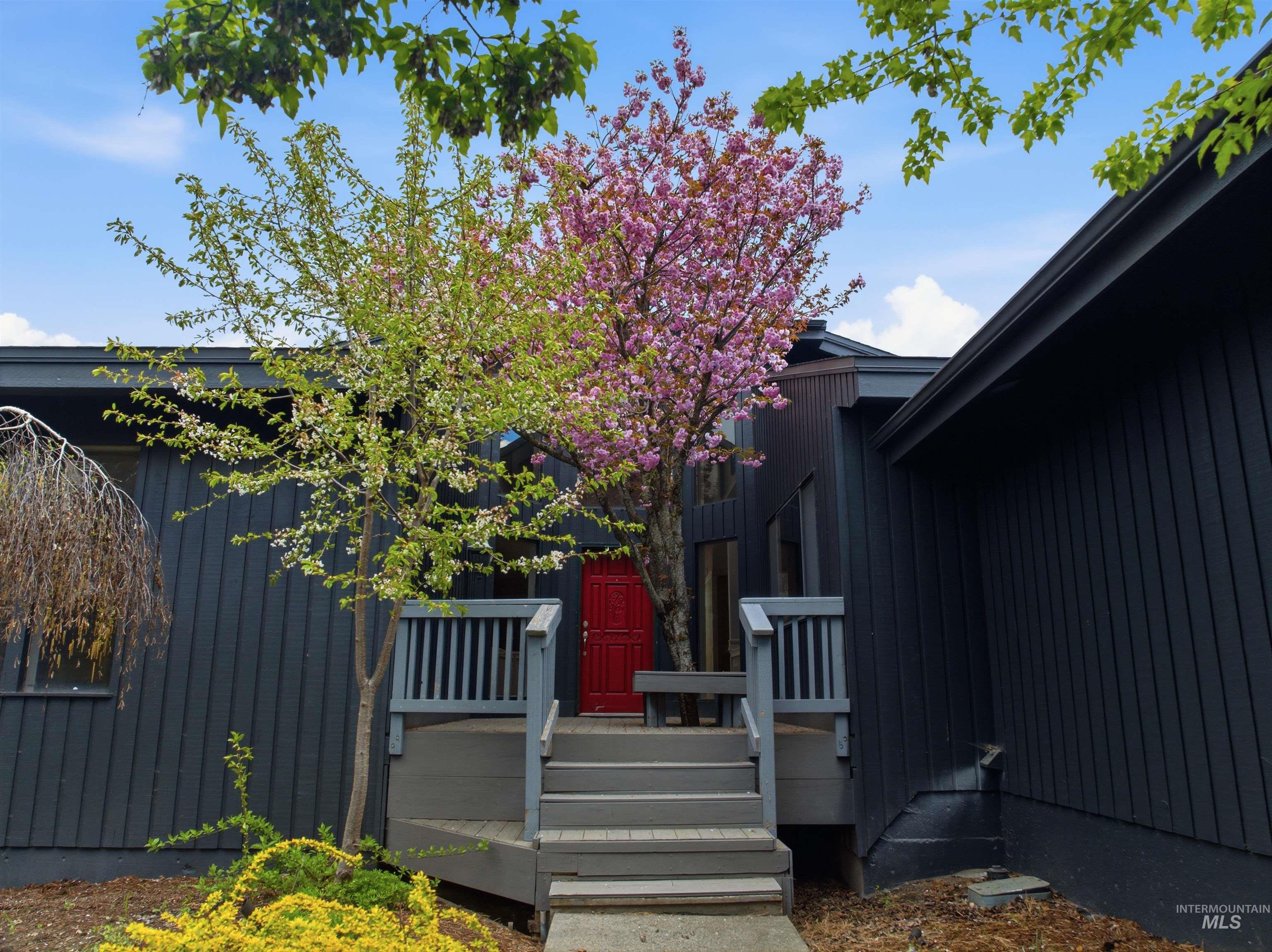 1540 Pine Cone Road Moscow, ID 83843 - Photo 3 of 49 Contemporary exterior featuring vertical siding, a striking red entry door, a multi-tiered deck with railings, and mature landscaping