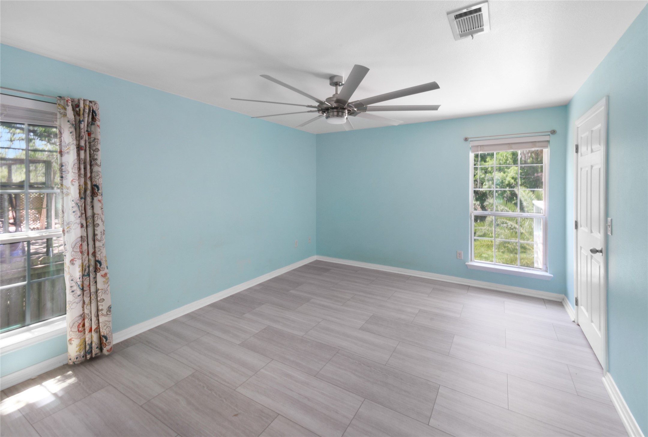 320 10th Street Dickinson, TX 77539 - Photo 18 of 21 Experience tranquility in this spacious primary bedroom, featuring soft blue walls, sleek tile flooring, and abundant natural light through large windows, complemented by a stylish ceiling fan for ultimate comfort.