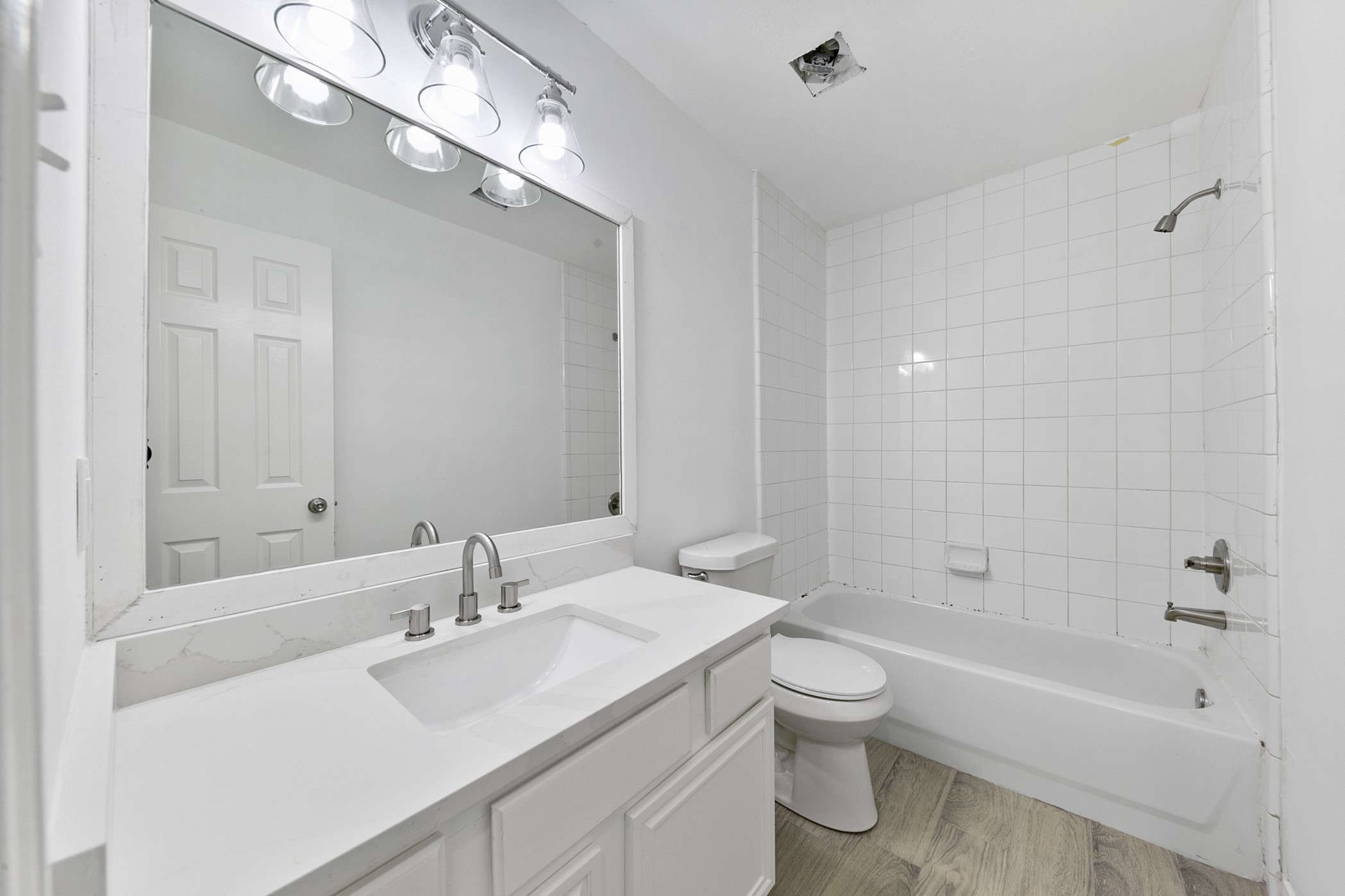 12014 Raven View Drive Houston, TX 77067 - Photo 24 of 29 This photo showcases a modern, bright bathroom featuring a sleek white vanity with a large mirror, overhead lighting, tiled shower-tub combo, and wood-style flooring.