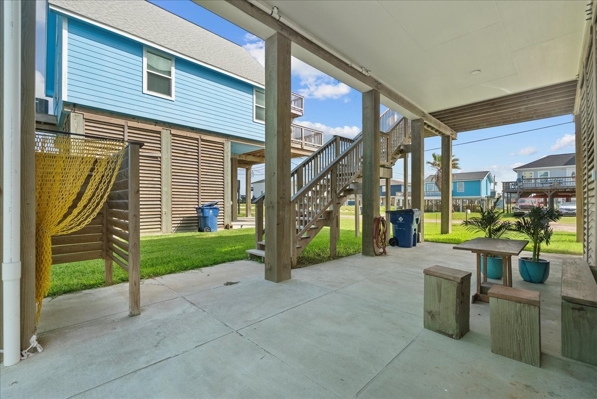 130 Beachcomber Avenue Surfside Beach, TX 77541 - Photo 28 of 33 This photo shows a covered outdoor area with a concrete floor, featuring a small wooden picnic table and potted plants. There's a yellow hammock on the left, and stairs lead up to a raised house, with grassy surroundings and neighboring homes in view.