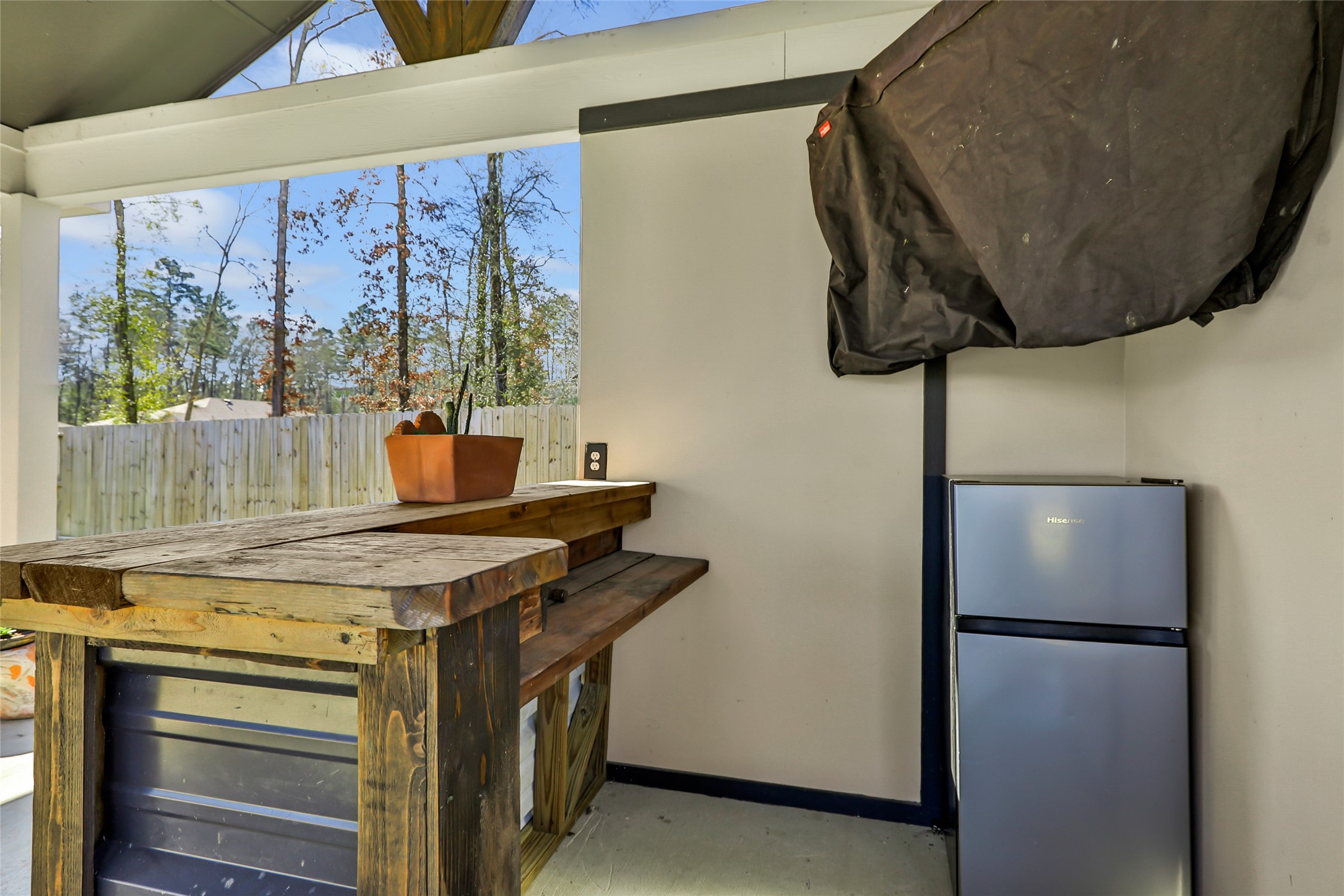 811 Republic Road New Caney, TX 77357 - Photo 8 of 37 This photo shows a cozy outdoor space featuring a rustic wooden bar, a small fridge, and a grill covered with a protective cover. The area is partially enclosed with a view of a fenced yard and trees, ideal for entertaining or relaxing.